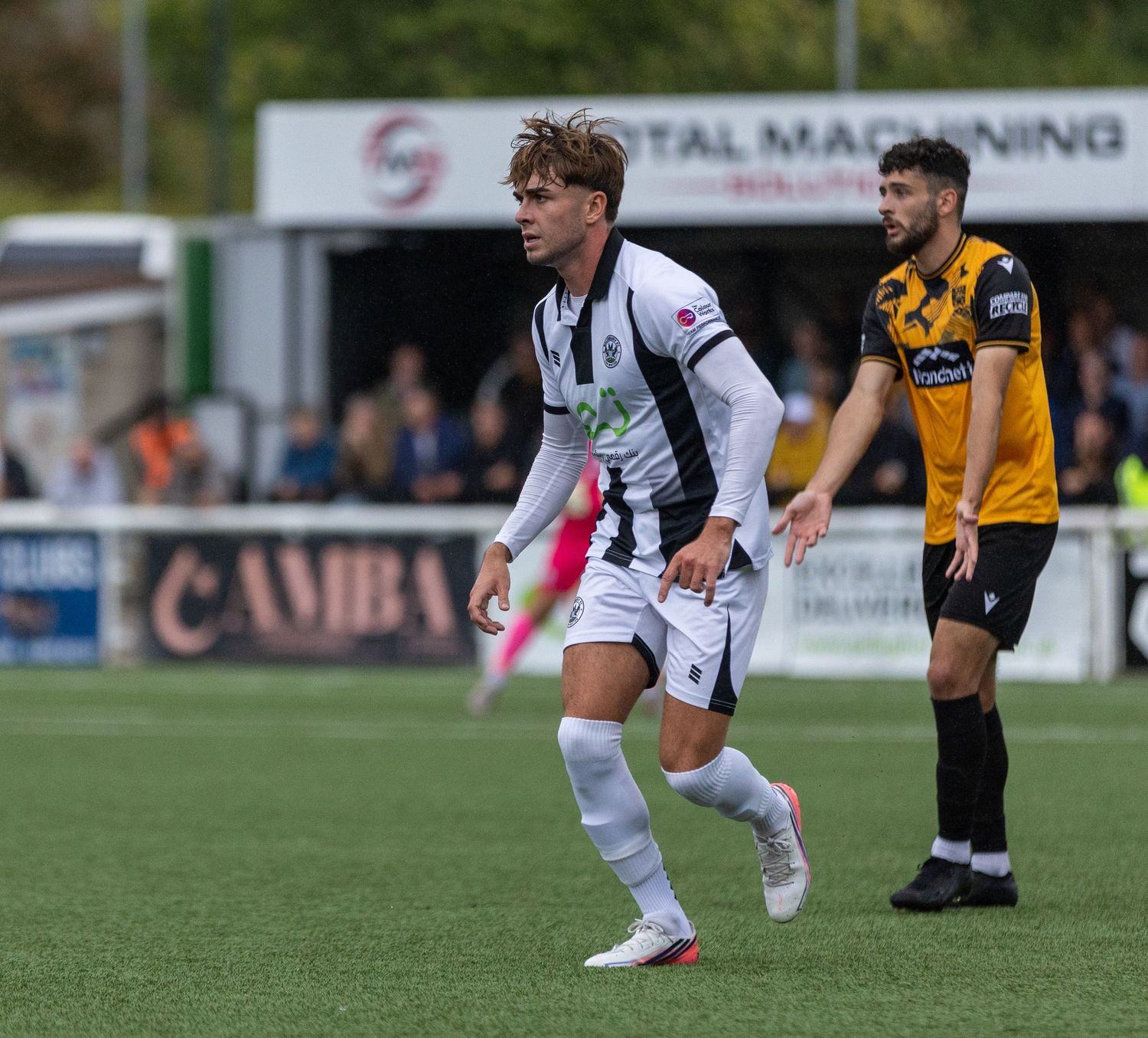 Two soccer players in action during a match, one in white and black kit, another in yellow and black, on a green pitch.