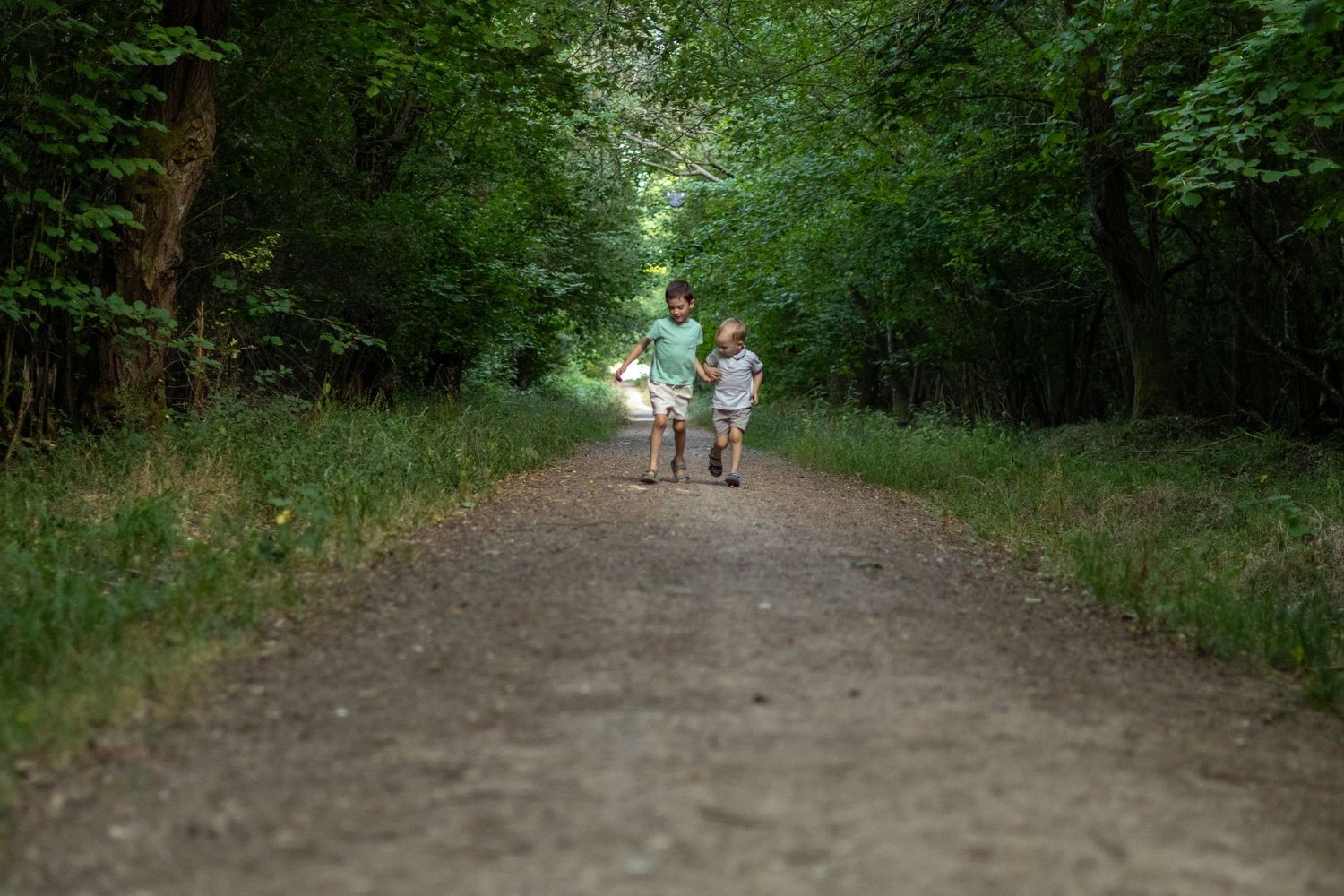 Two young children holding hands strolling along a peaceful dirt path in a lush green forest.