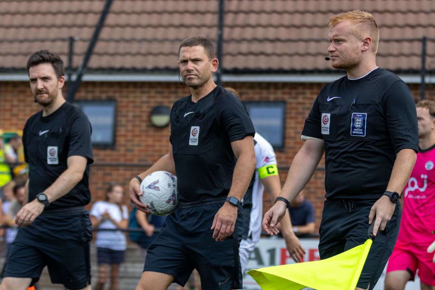 Three football referees dressed in black uniforms walk together onto a non-league soccer pitch before a match.