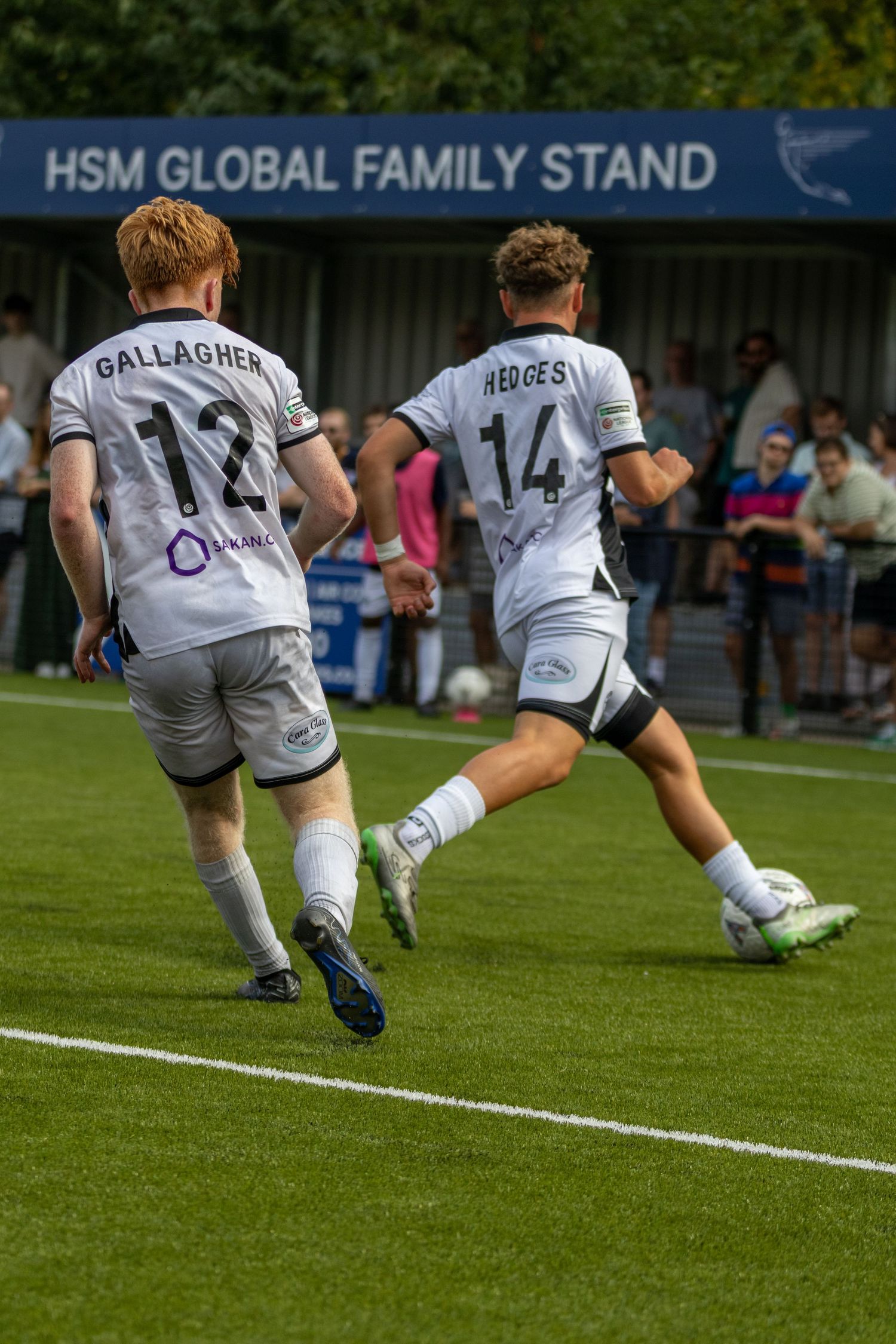Gallagher #12 and Hedges #14 in white kits play near the HSM Global Family Stand during a football match.