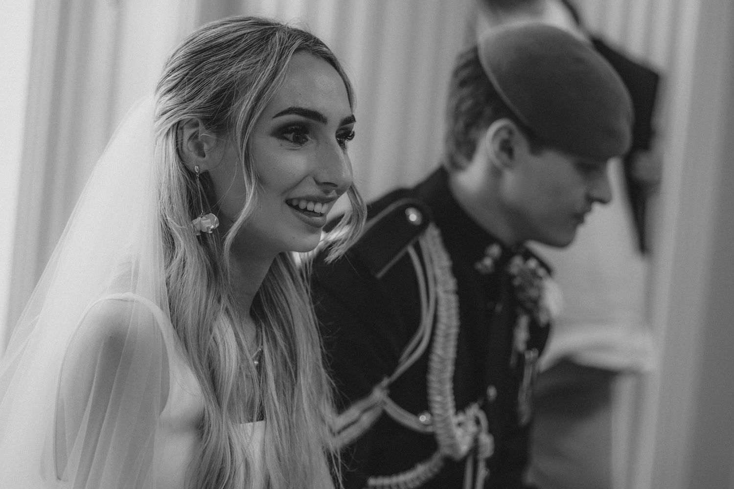 Black and white photo of smiling blonde bride in veil beside military officer at wedding ceremony.