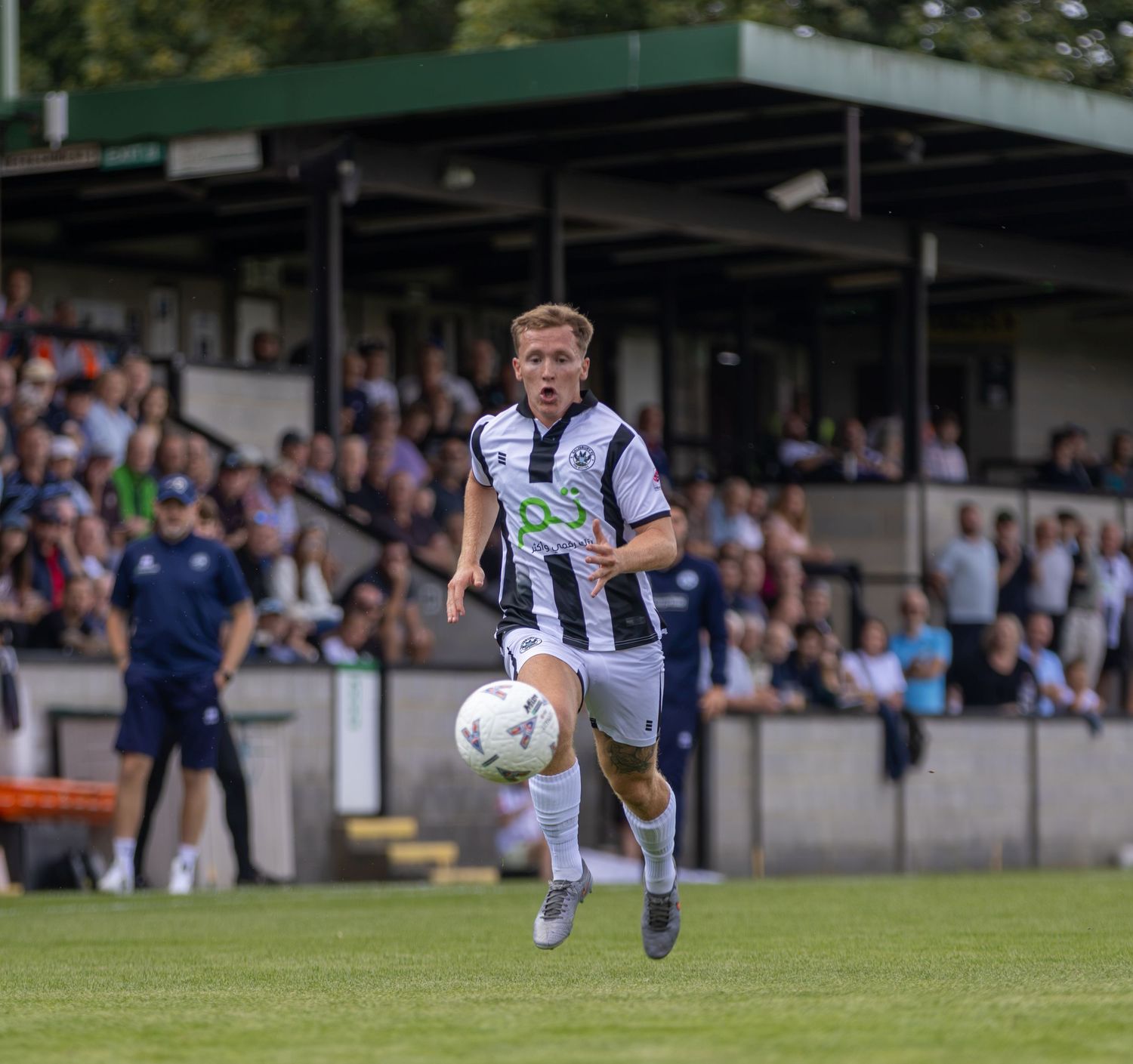 Blonde footballer in black and white striped kit controls the ball while running on a pitch with packed stands behind.