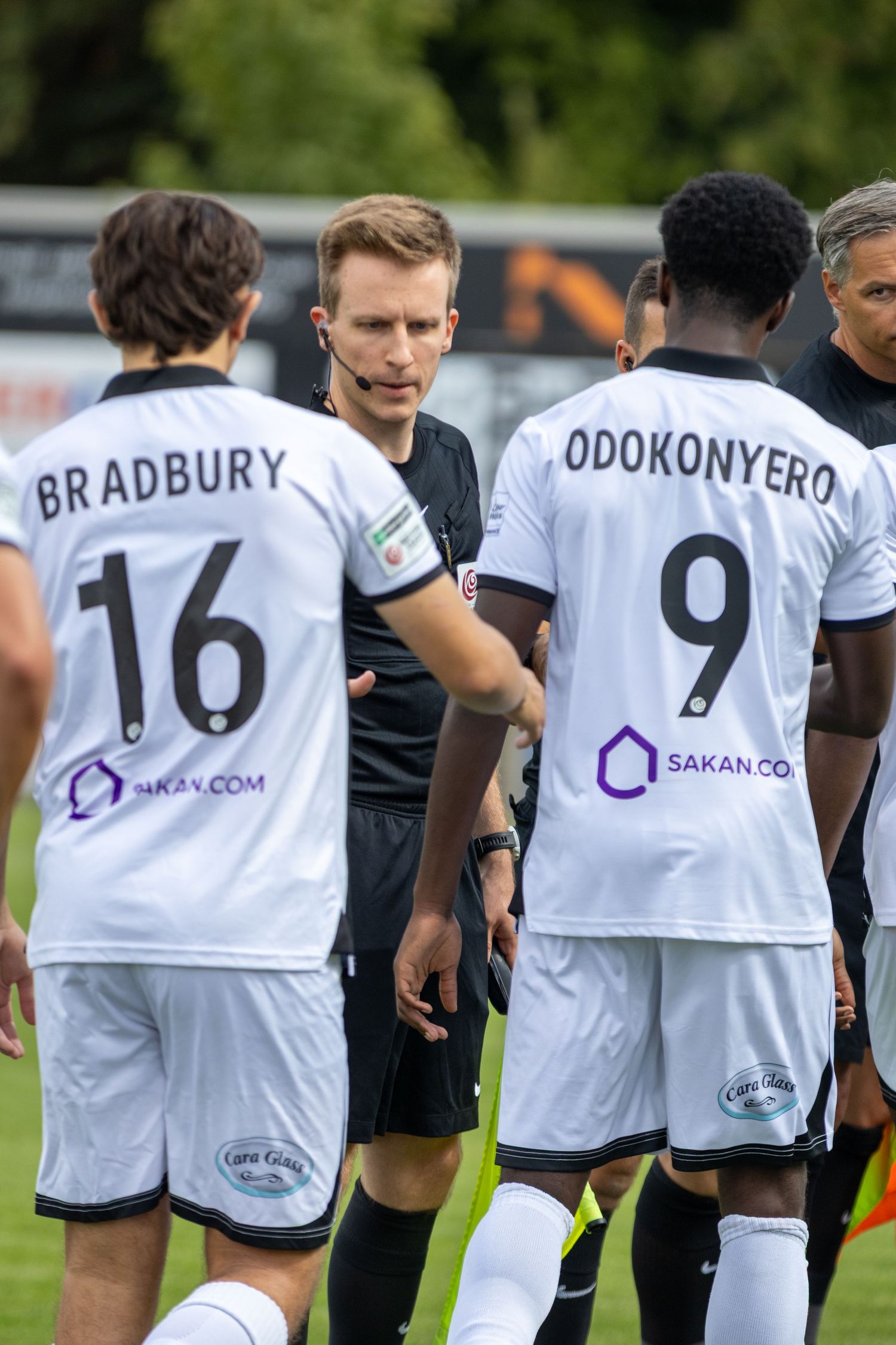 Football referee addressing players Bradbury 16 and Odokonyero 9 wearing white Sakan.com sponsored kits.