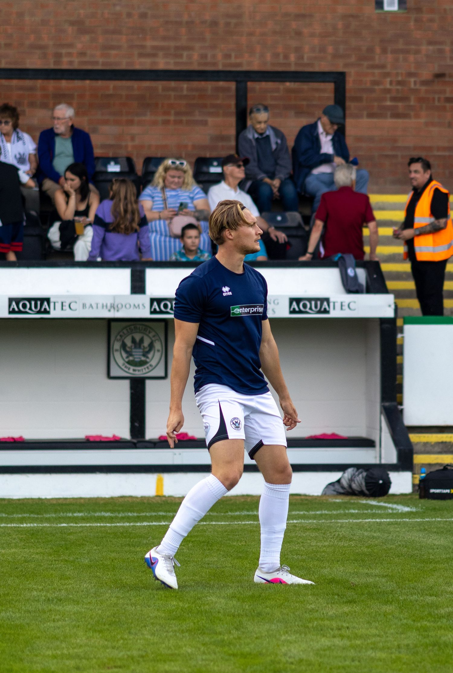 A blond footballer in navy Errea kit walks across the pitch during a football match at a stadium.