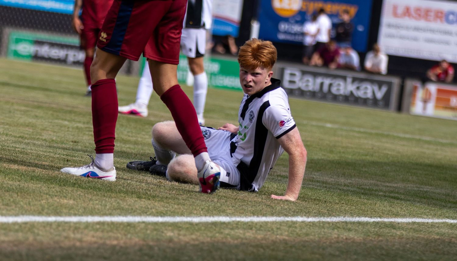 A red-haired soccer player in black and white kit sits on the grass during a sunny football match.