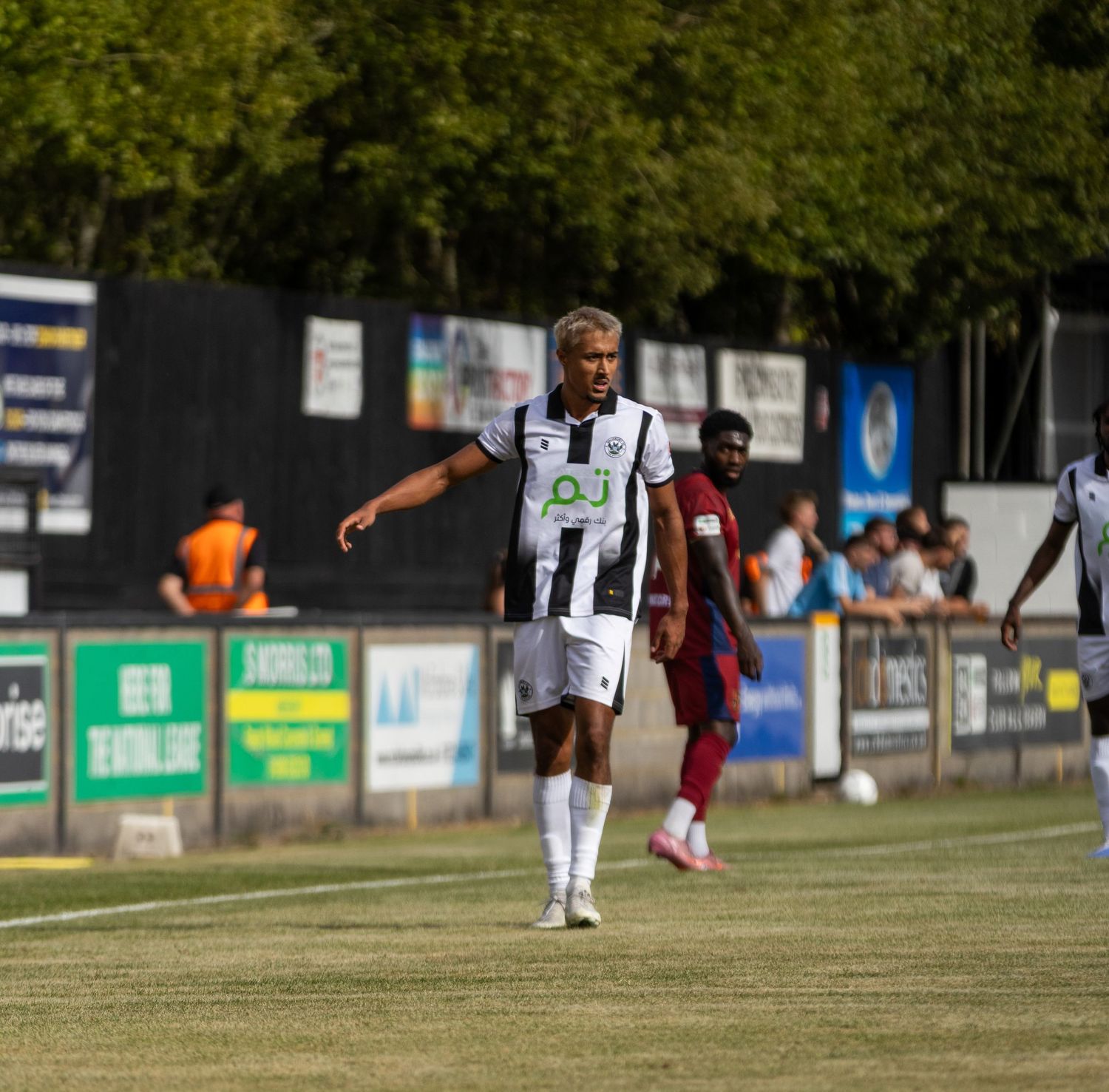 A footballer in black and white kit walks along the touchline during a sunny non-league match.