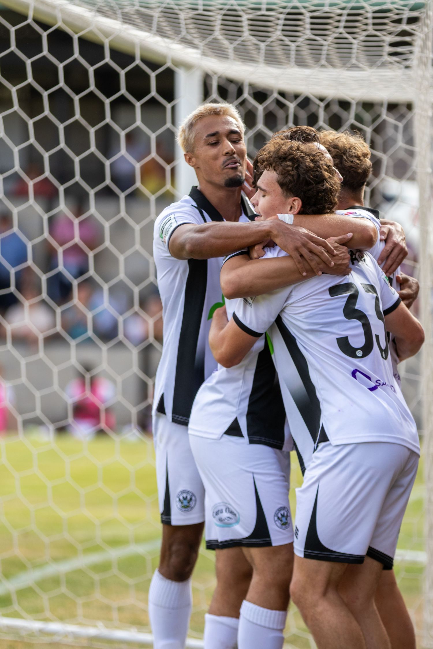 Three soccer players in black and white uniforms share a celebratory hug near the goal.
