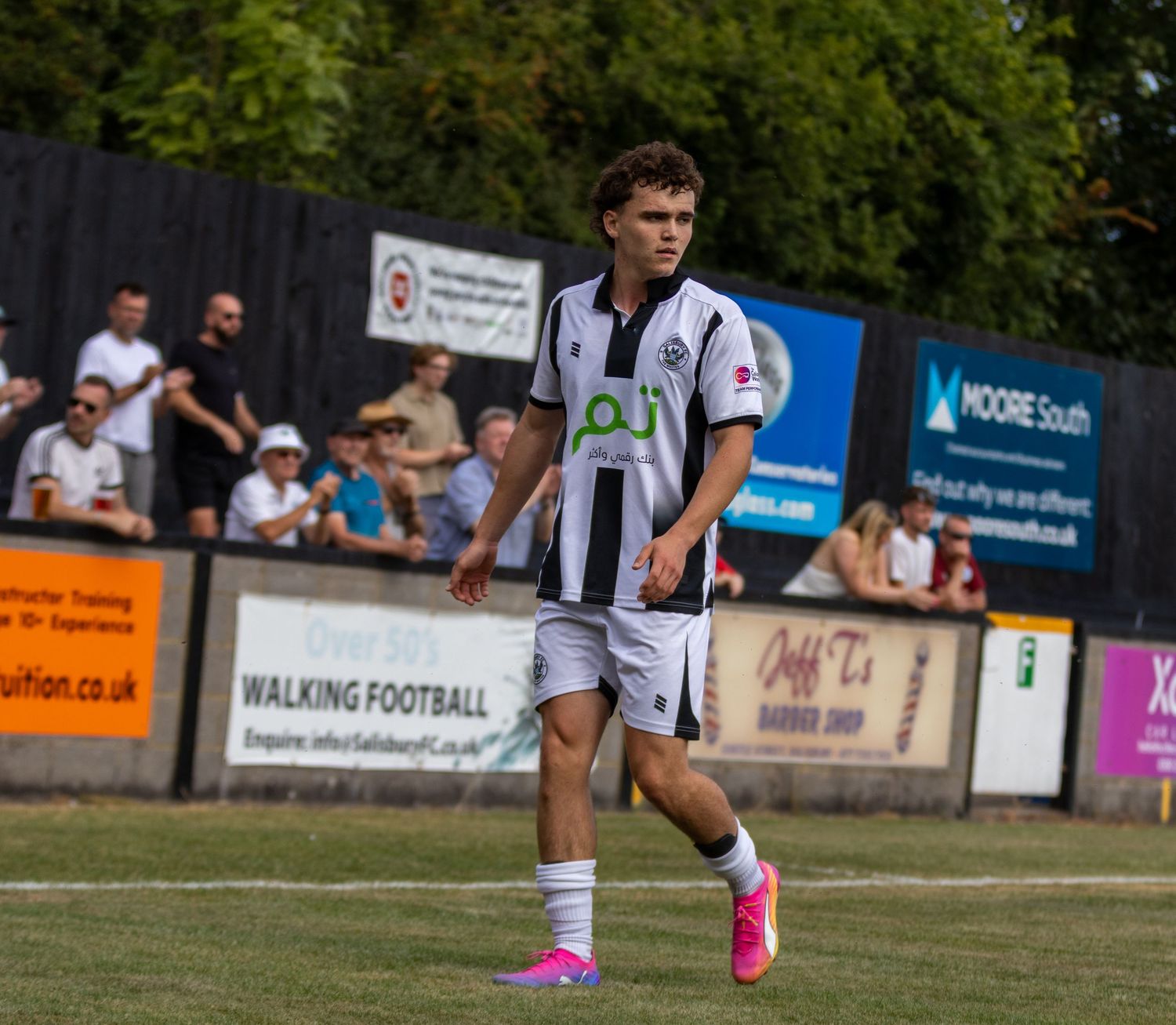 A young male footballer in black and white striped kit walks across the pitch during a sunny match.