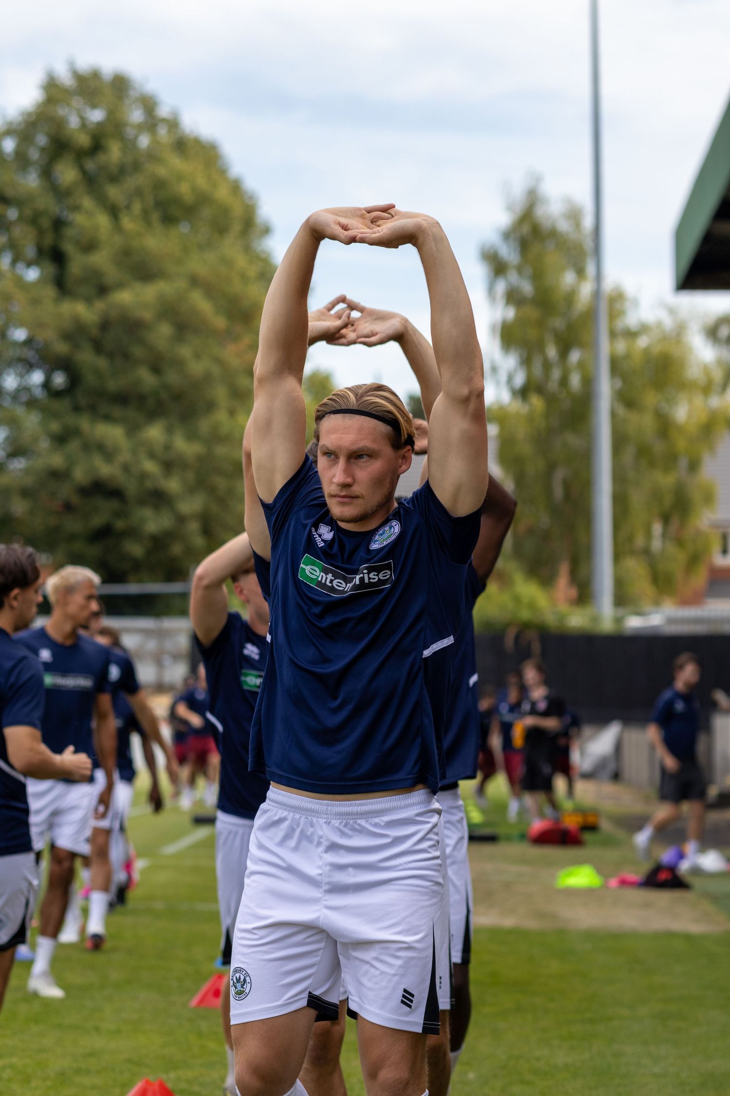 Football player in navy Enterprise kit stretching arms overhead during outdoor training session warmup.