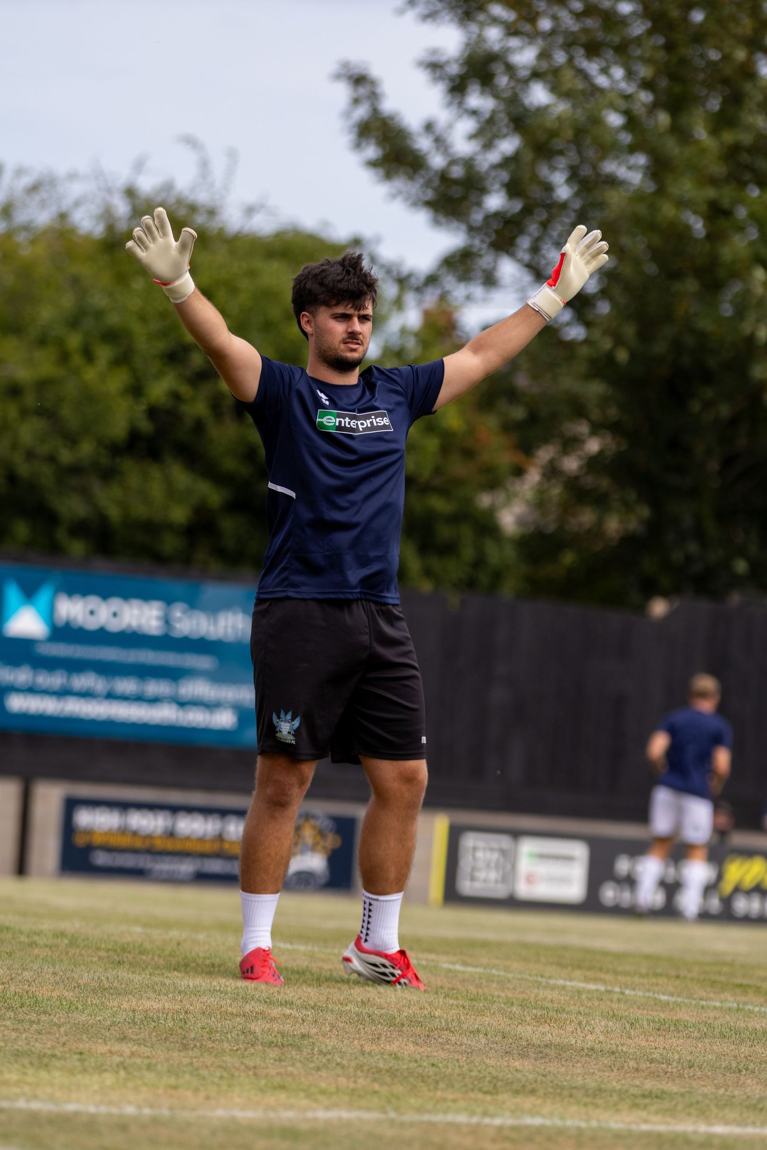 A goalkeeper in navy blue kit raises both gloved hands on a grass football pitch during a sunny match day.
