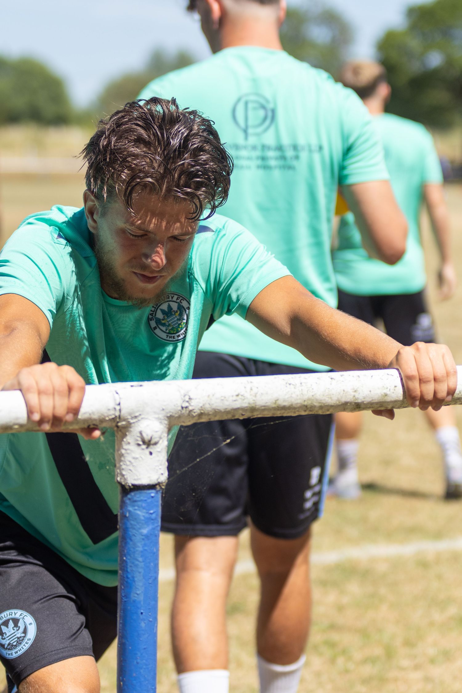 Sweating footballer in teal kit pushing weighted bar during intense summer training session.