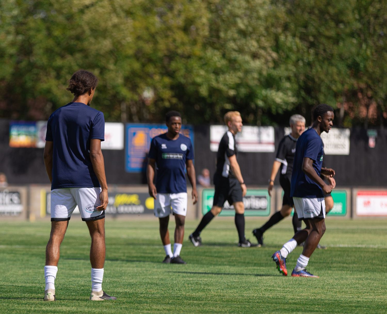 Soccer players in navy uniforms warm up on a green field during a sunny outdoor match or practice session.
