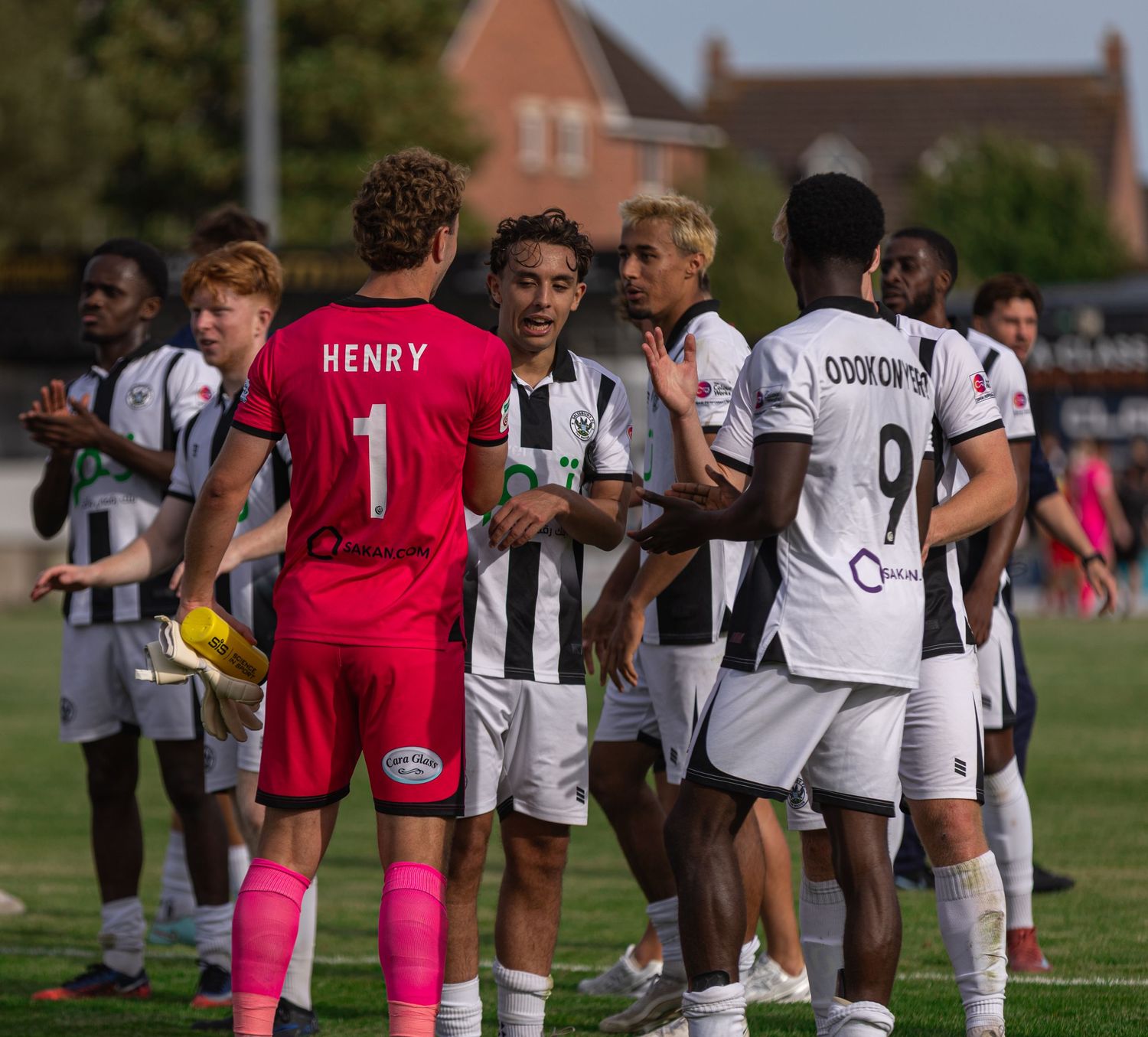 Soccer players in black and white uniforms, including goalkeeper Henry wearing number 1, gather on a sunny outdoor pitch.
