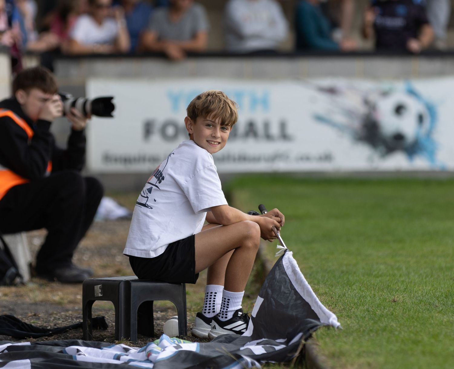 A smiling young boy sits on a bench at a youth football event, wearing a white t-shirt and shorts on a sunny day.