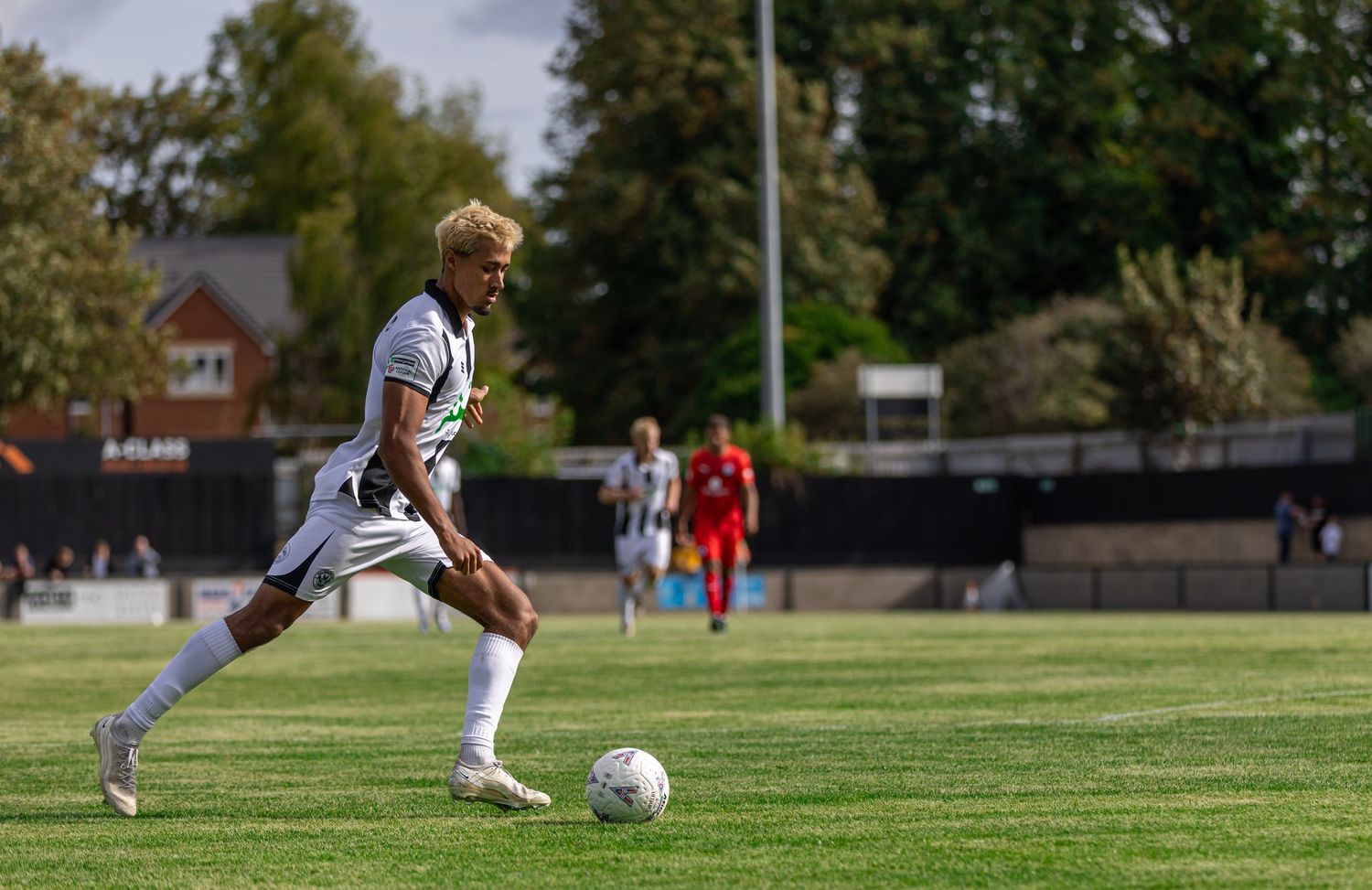 Soccer player in white kit dribbling a ball on a green pitch during a sunny outdoor match.