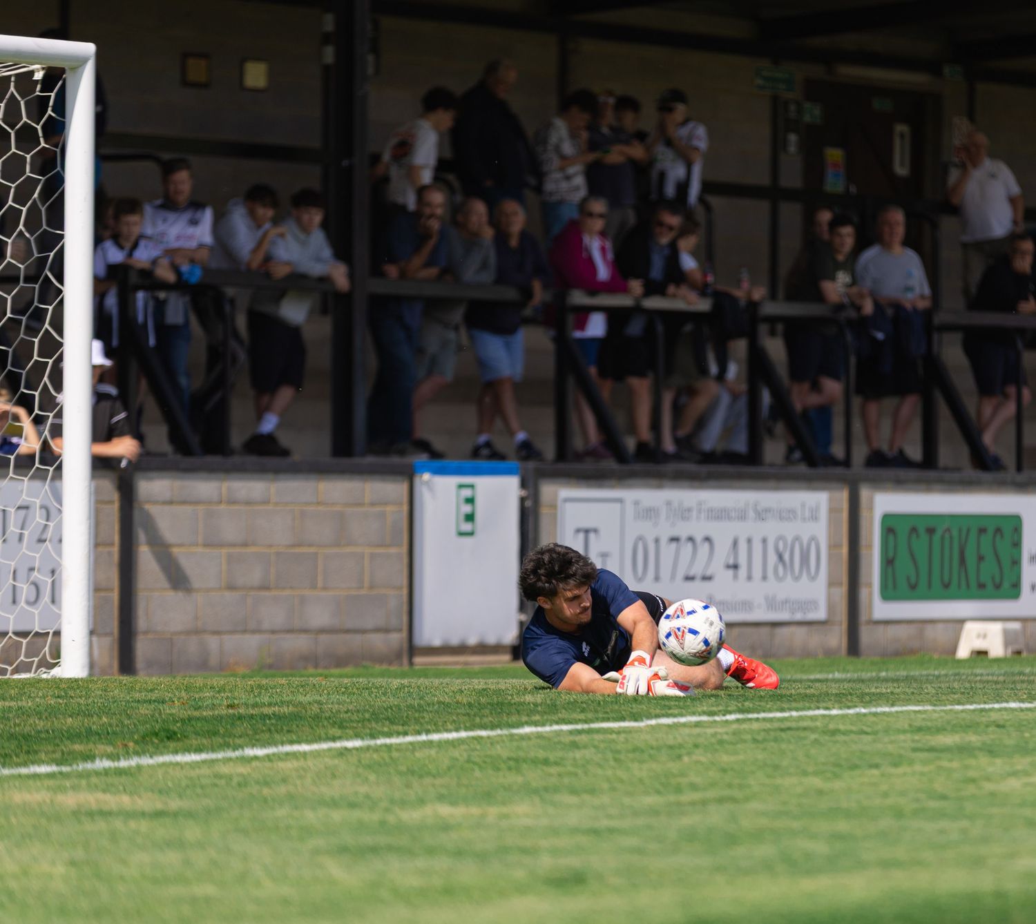 Goalkeeper diving to save a ball during a football match, with spectators watching from the stands behind.