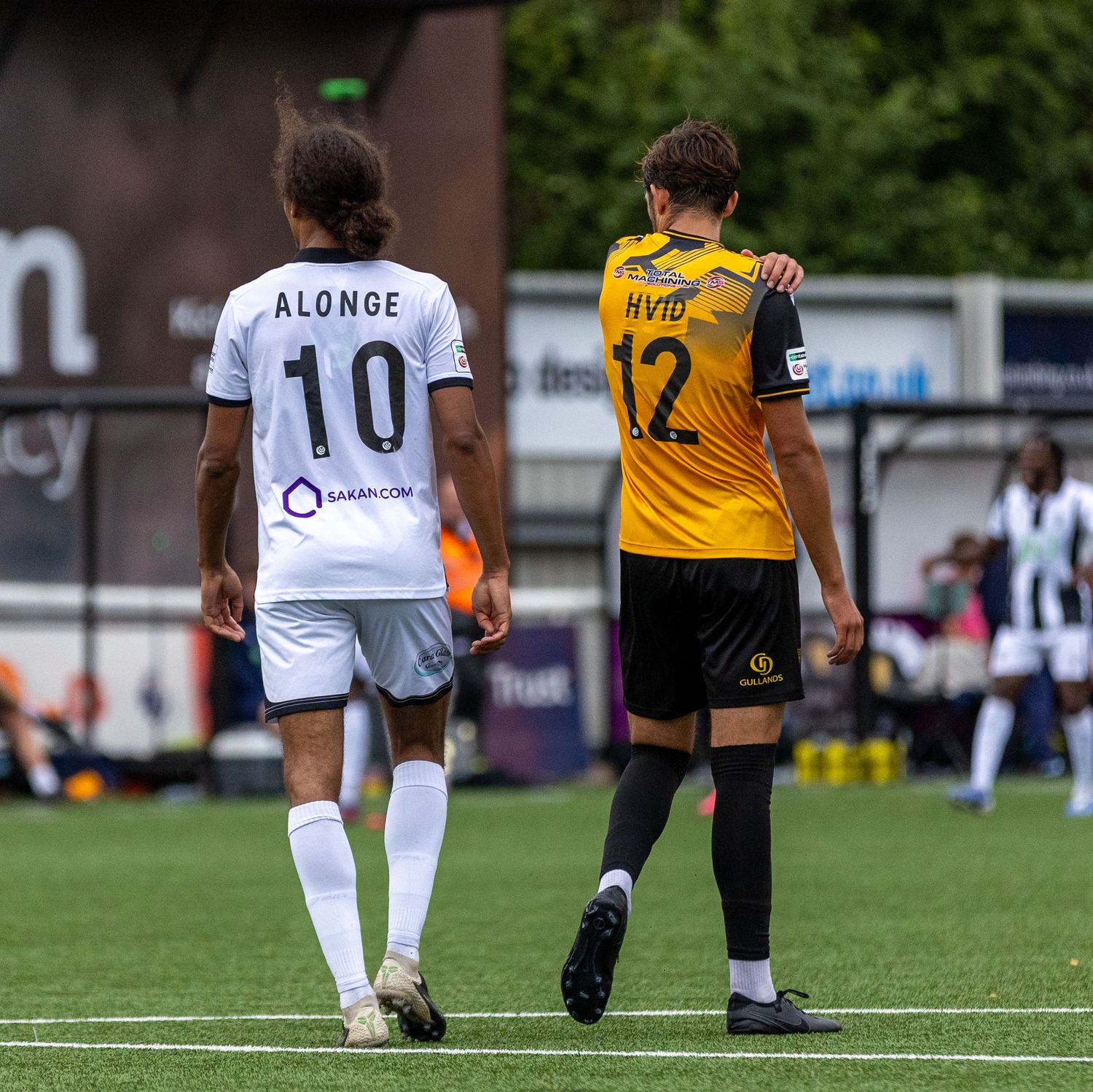 Two soccer players, Alonge #10 and Hvid #12, walk side by side on a green pitch during a match.