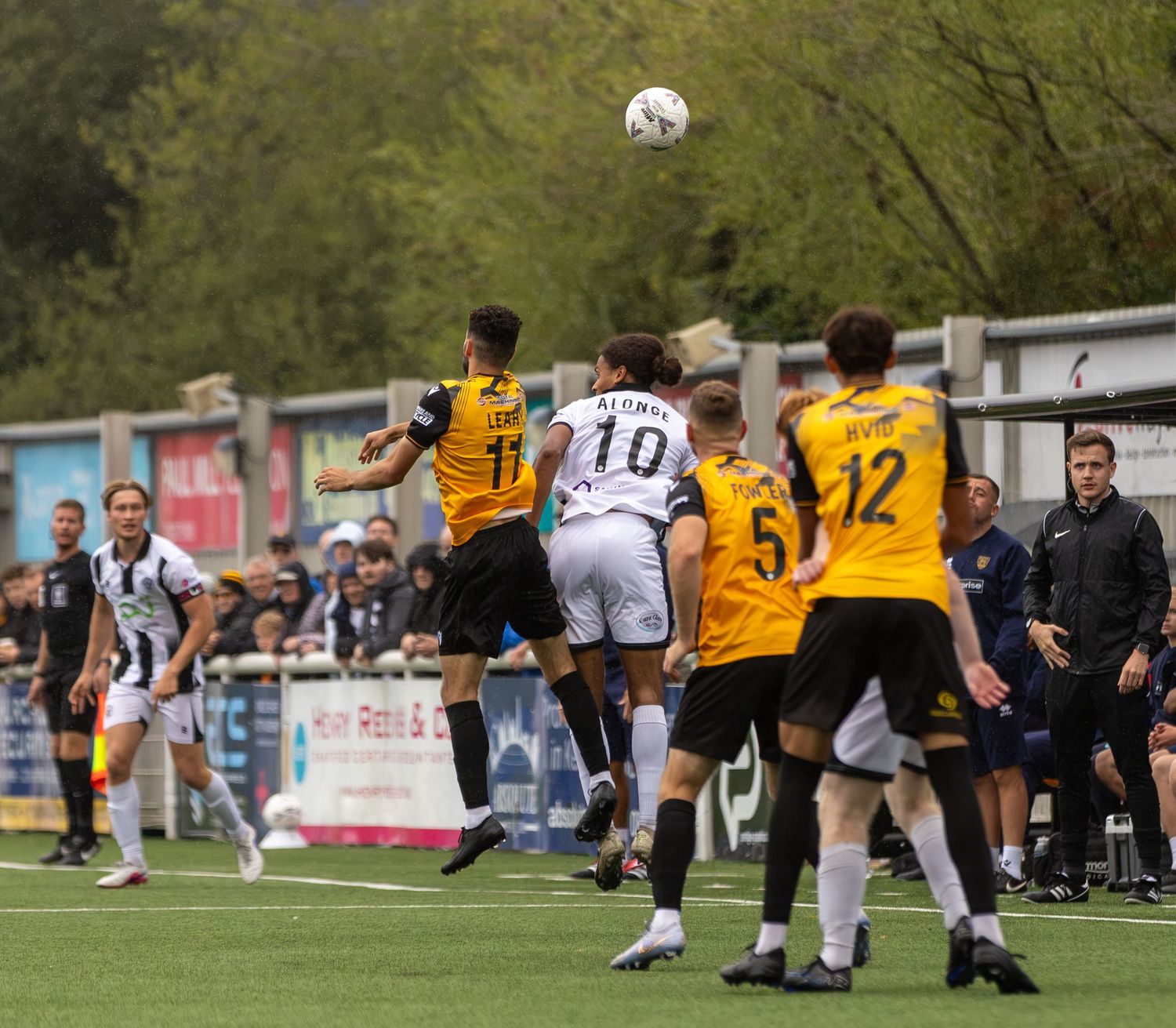 Soccer players compete for a header during an outdoor match, with fans watching in the background on a cloudy day.