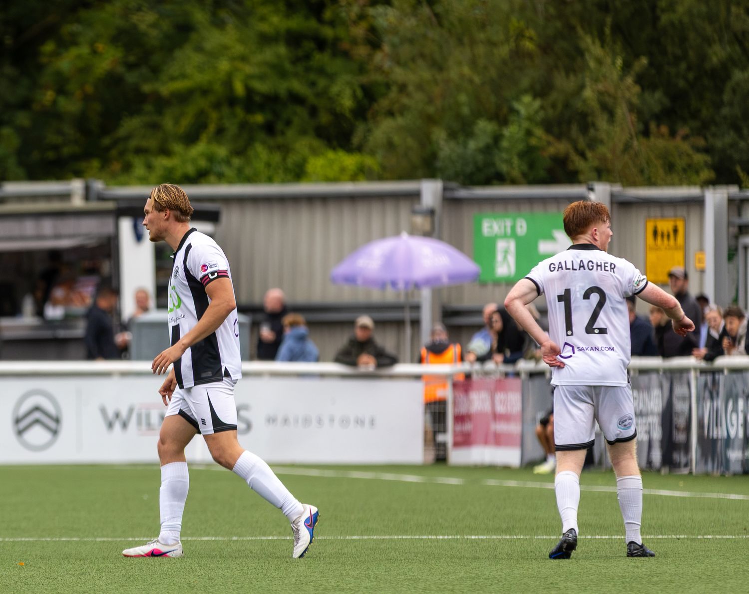 Two football players in black and white uniforms on a green pitch, number 12 Gallagher visible, with spectators in background.