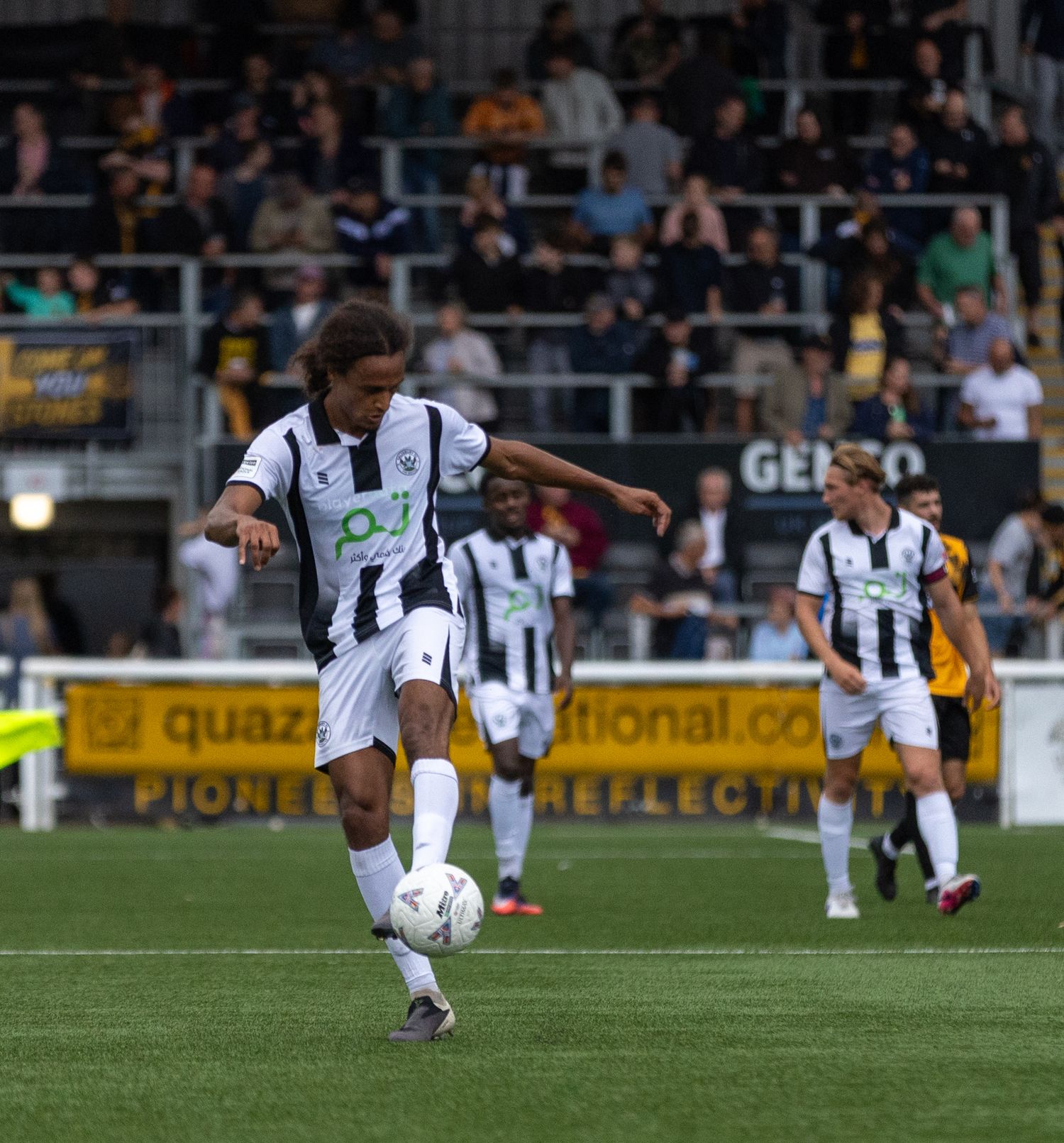 A soccer player in black and white uniform dribbles the ball during a competitive match in a crowded stadium.