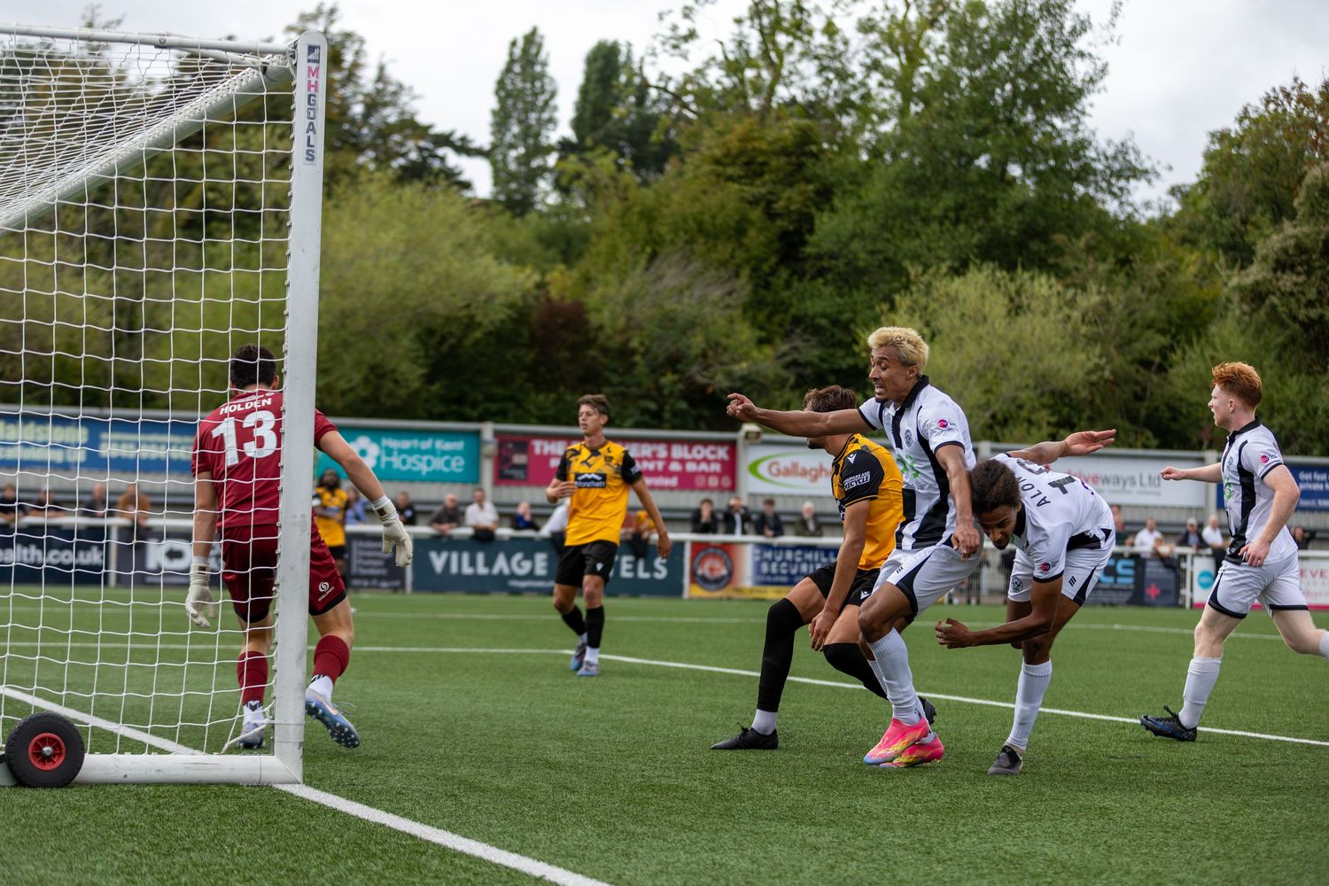 Soccer players compete near the goal during an outdoor match, with a goalkeeper in red and players in yellow and white.