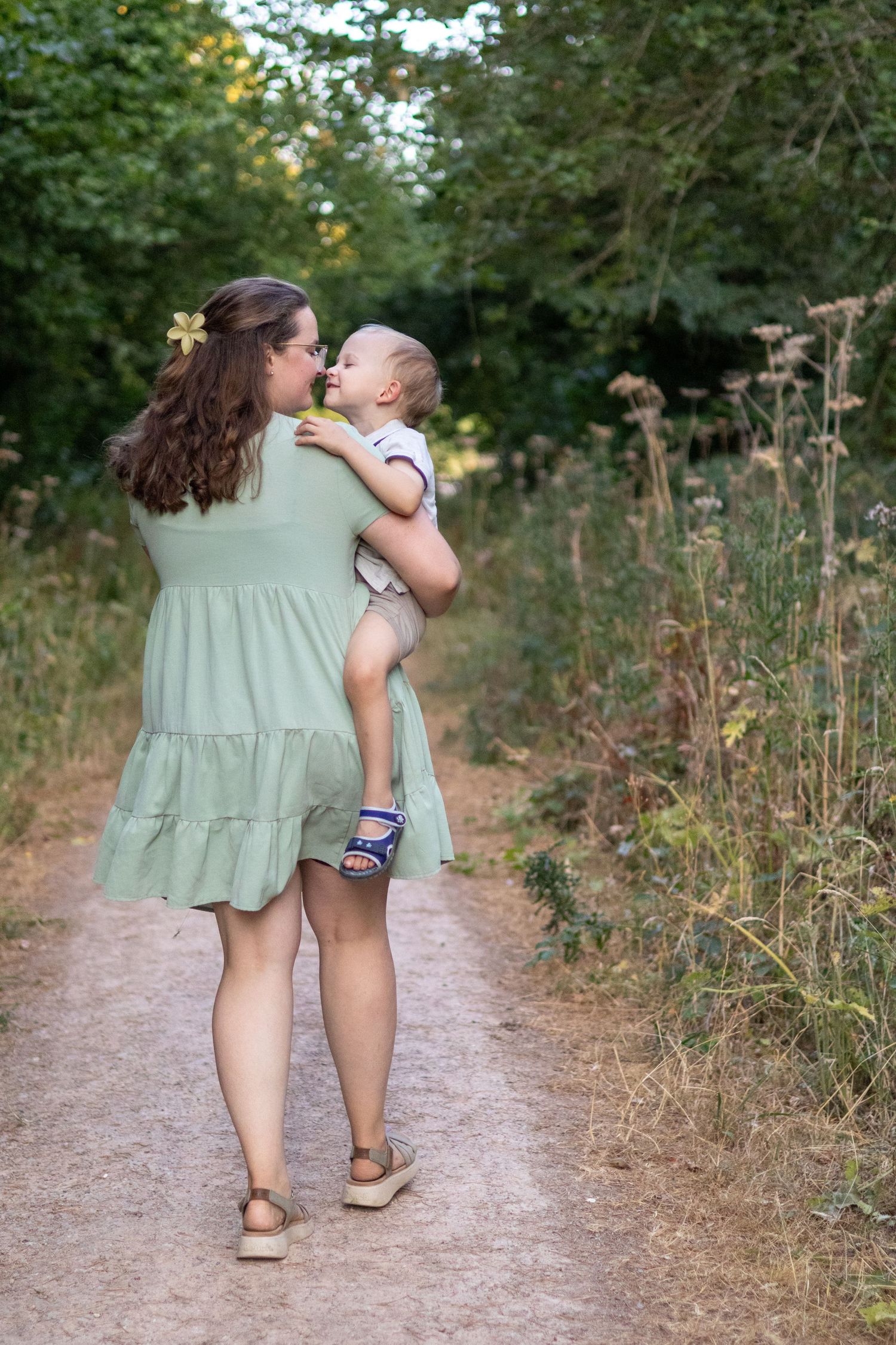 A mother in a sage green dress holds her baby on a nature path, sharing a tender nose-to-nose moment.