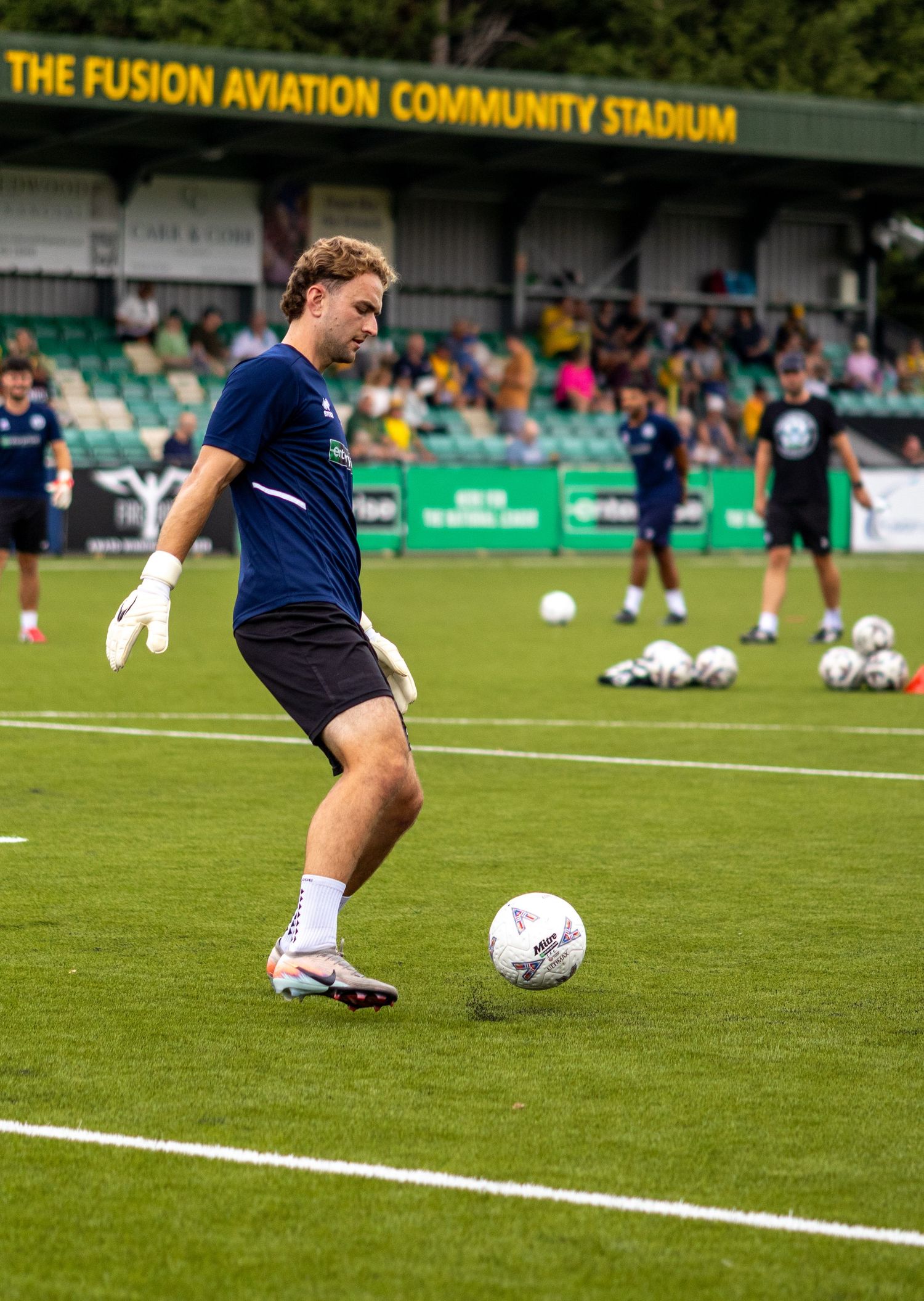 Goalkeeper in navy kit practicing ball control during pre-match warmup at Fusion Aviation Community Stadium.