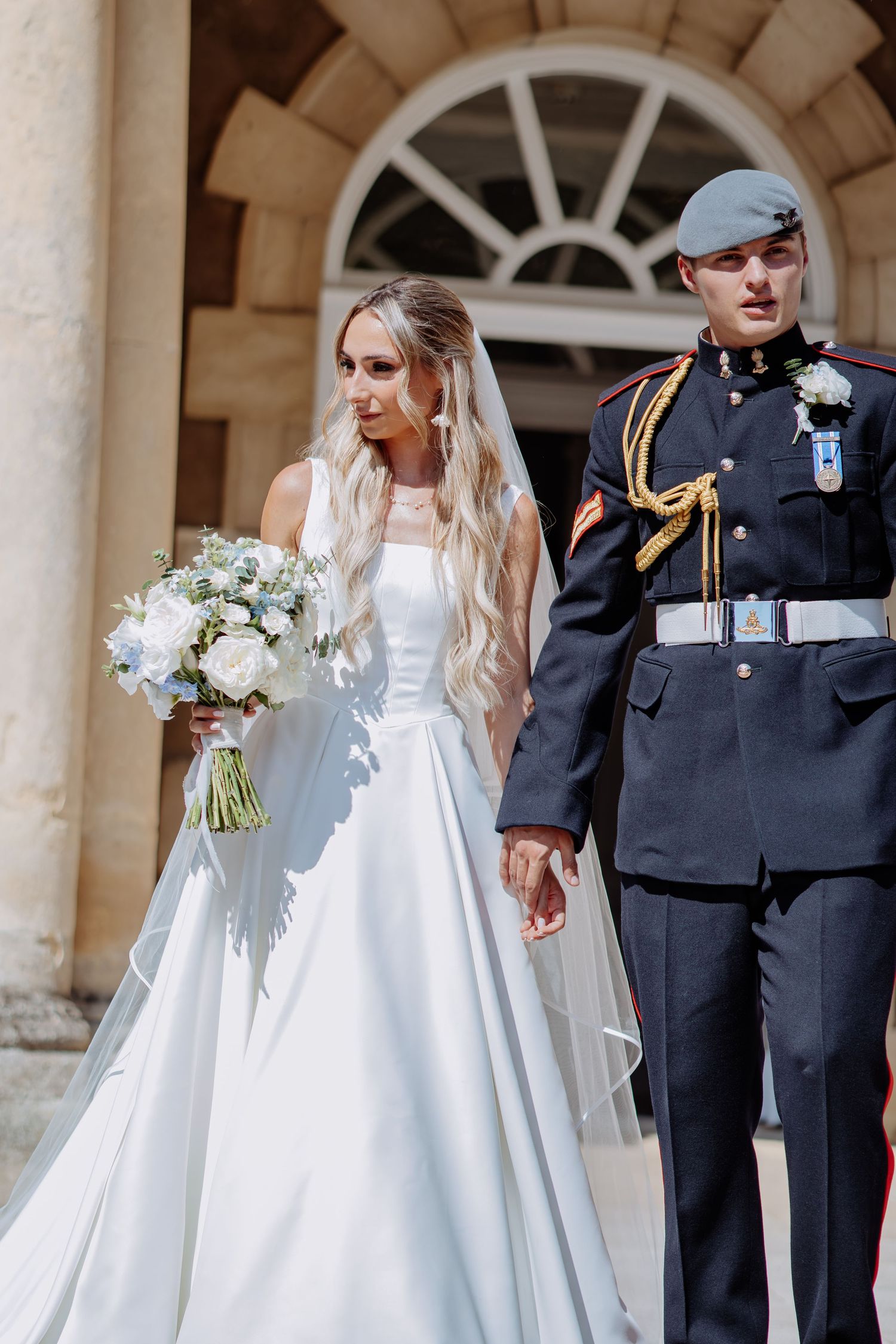Bride in white gown holding blue bouquet walks hand-in-hand with groom in military dress uniform outside elegant venue.
