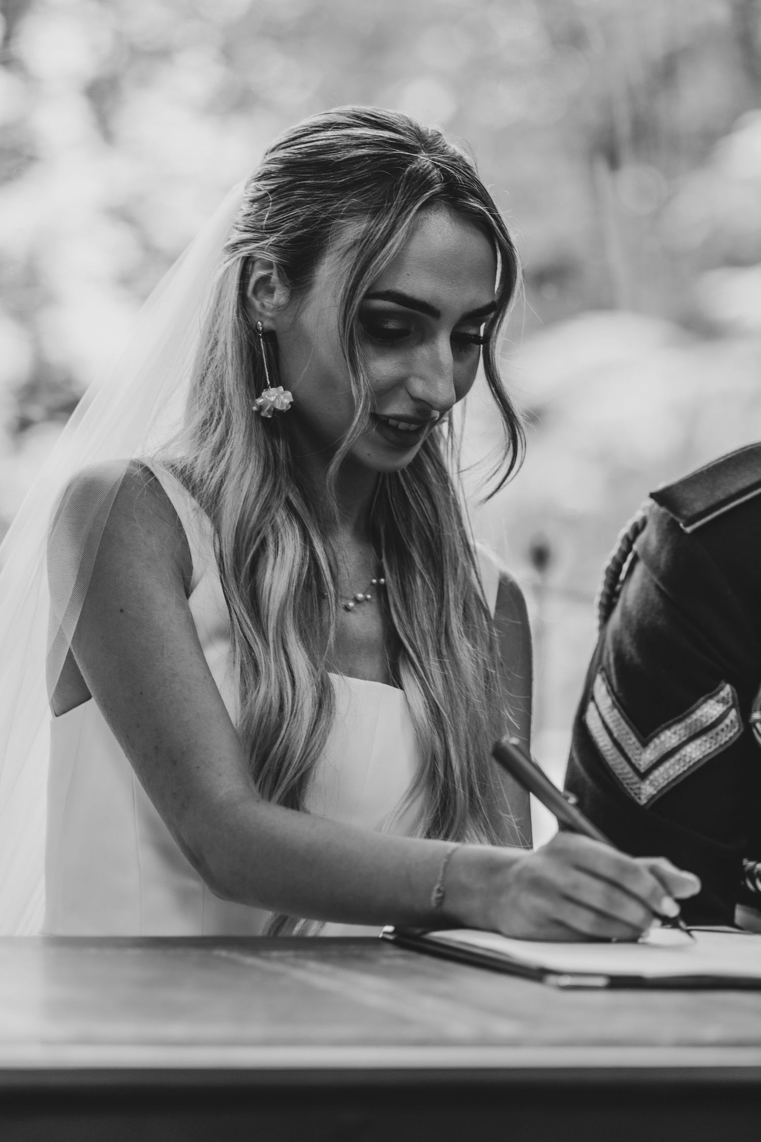 Black and white photo of blonde bride in veil signing wedding register at ceremony table.