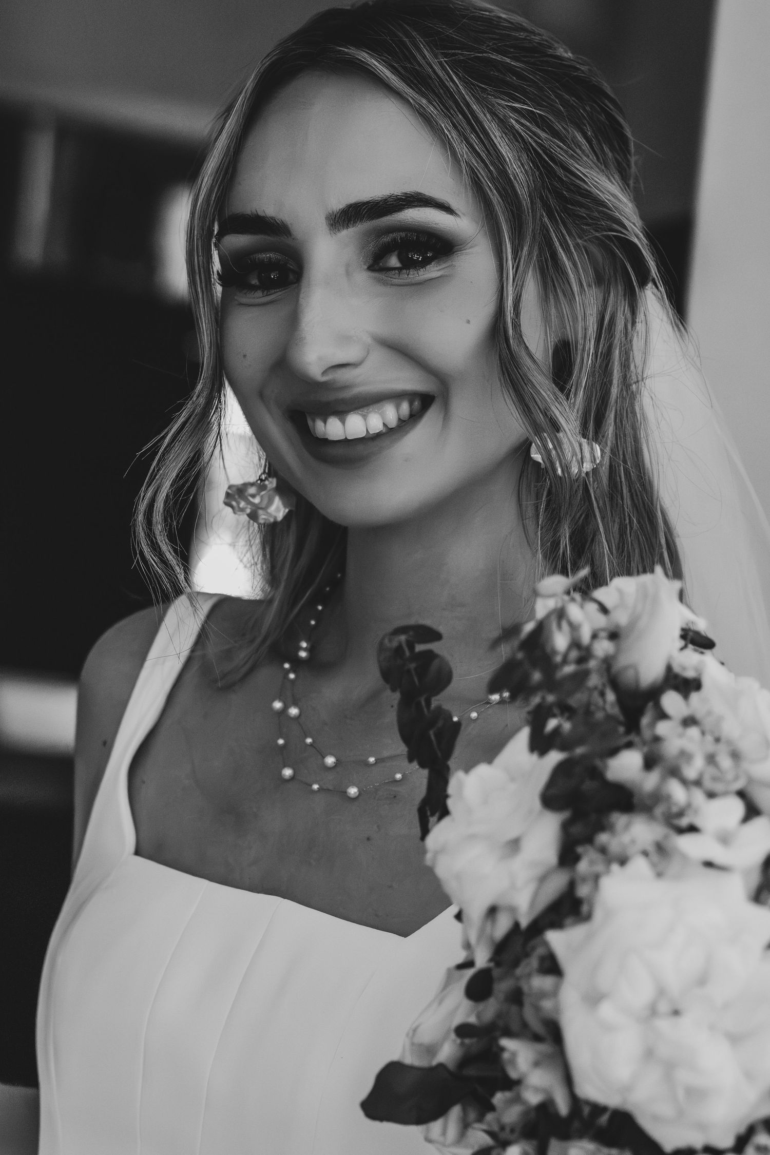 Black and white portrait of smiling bride wearing veil and pearl necklace holding white floral bouquet.