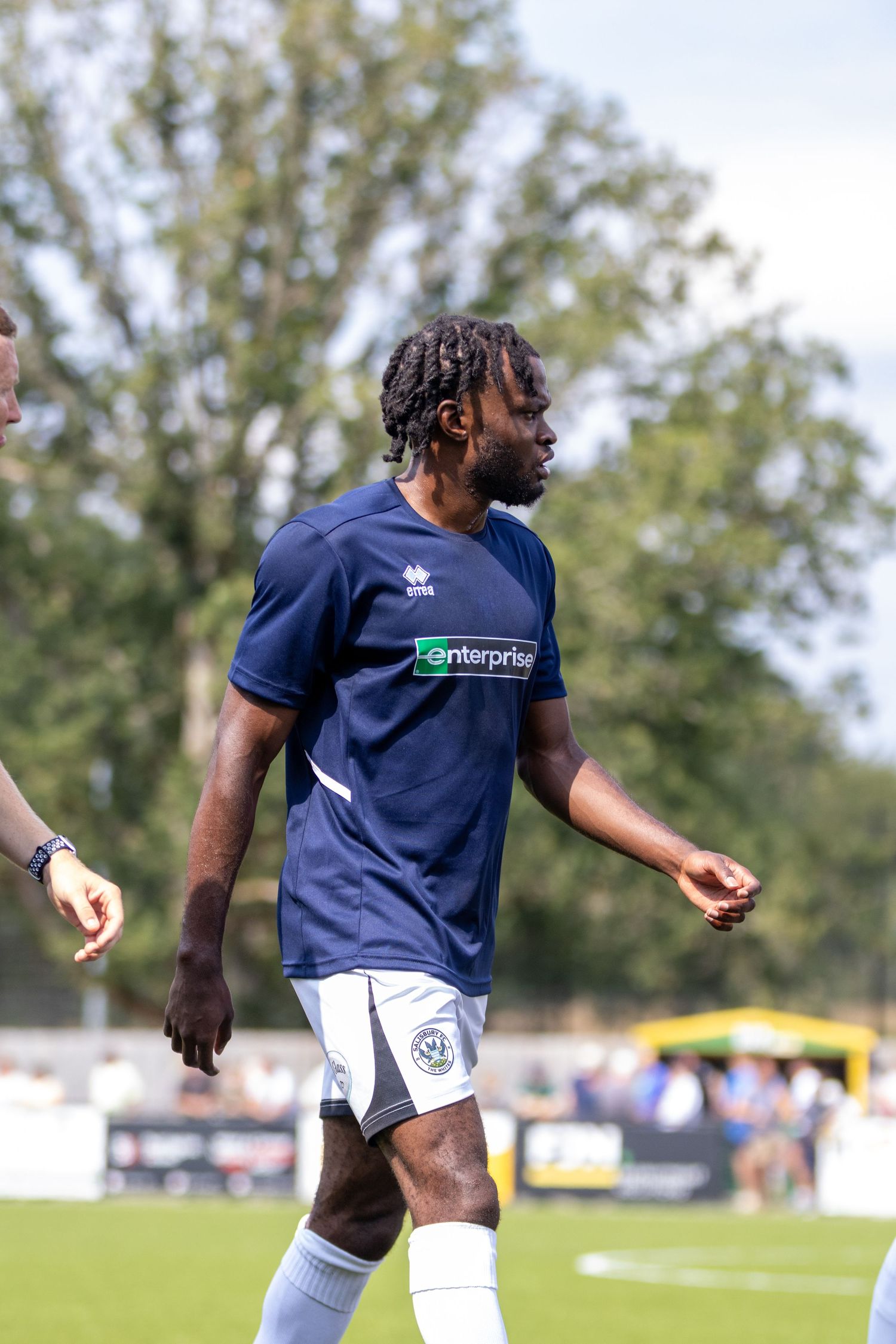 Male footballer with dreadlocks wearing navy Enterprise-sponsored kit walks across pitch during match.