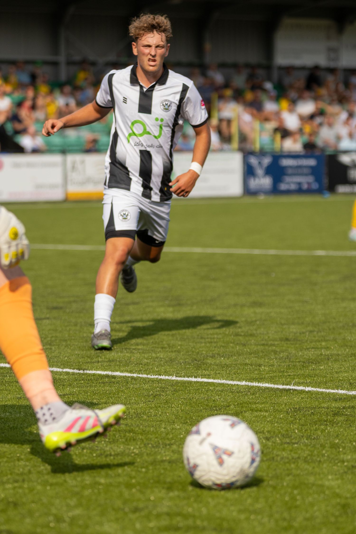 A curly-haired footballer in black and white striped kit chases the ball during a competitive outdoor match.