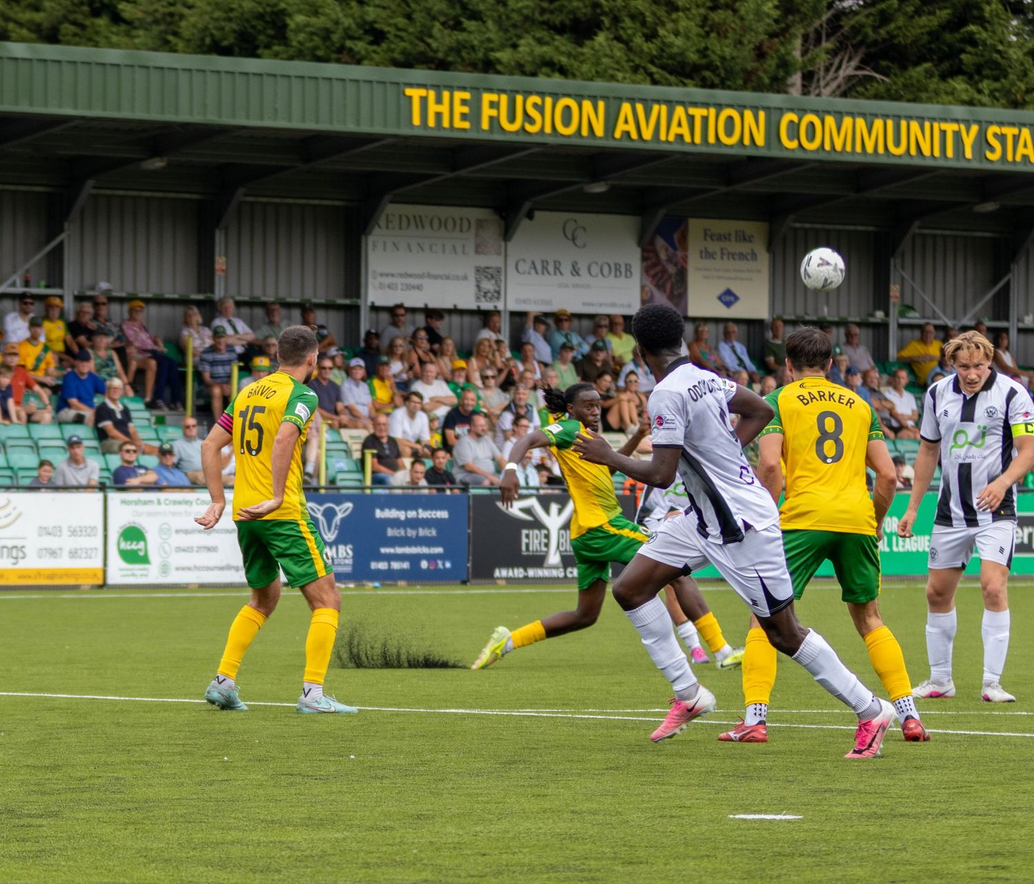 Footballers in yellow-green and black-white kits compete for a header at Fusion Aviation Community Stadium.
