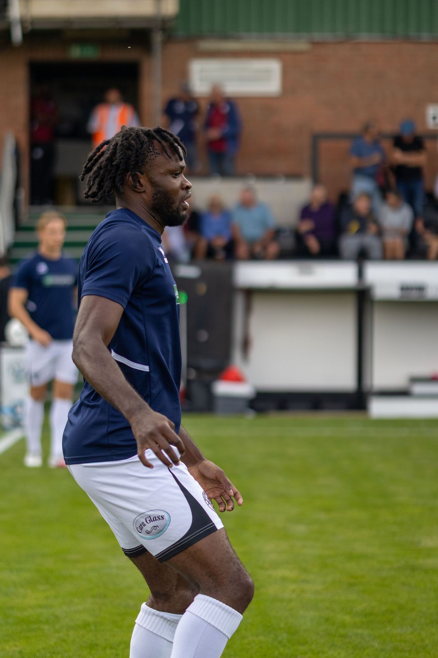 A Black male footballer with dreadlocks in a navy blue kit stands alert on a football pitch with fans behind.