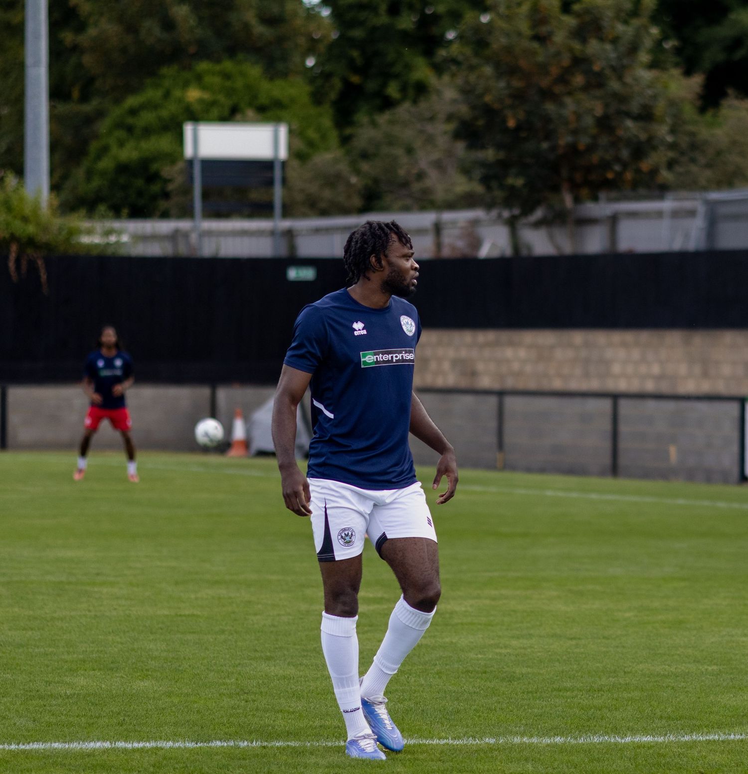 A footballer in navy blue Errea kit jogs across a green pitch during a pre-season football match.