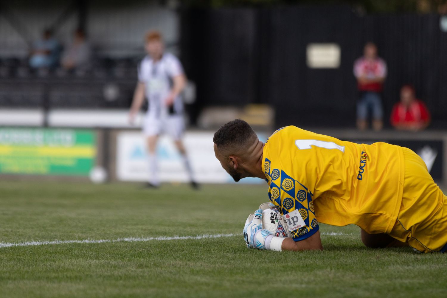 A goalkeeper in yellow kit dives to the ground to collect the ball during a football match.
