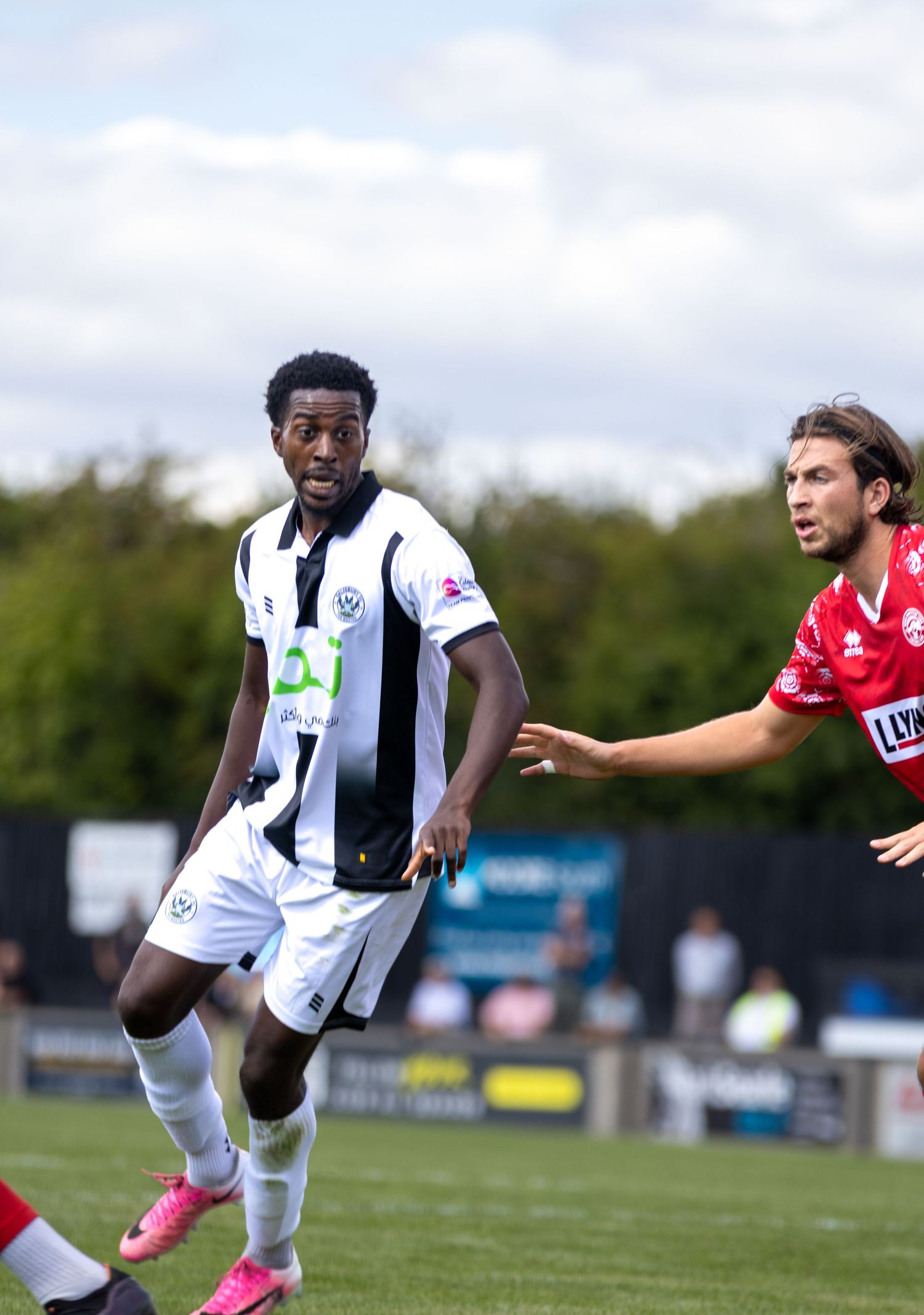 Footballer in black and white striped kit shields ball from red-kitted opponent under a partly cloudy sky.