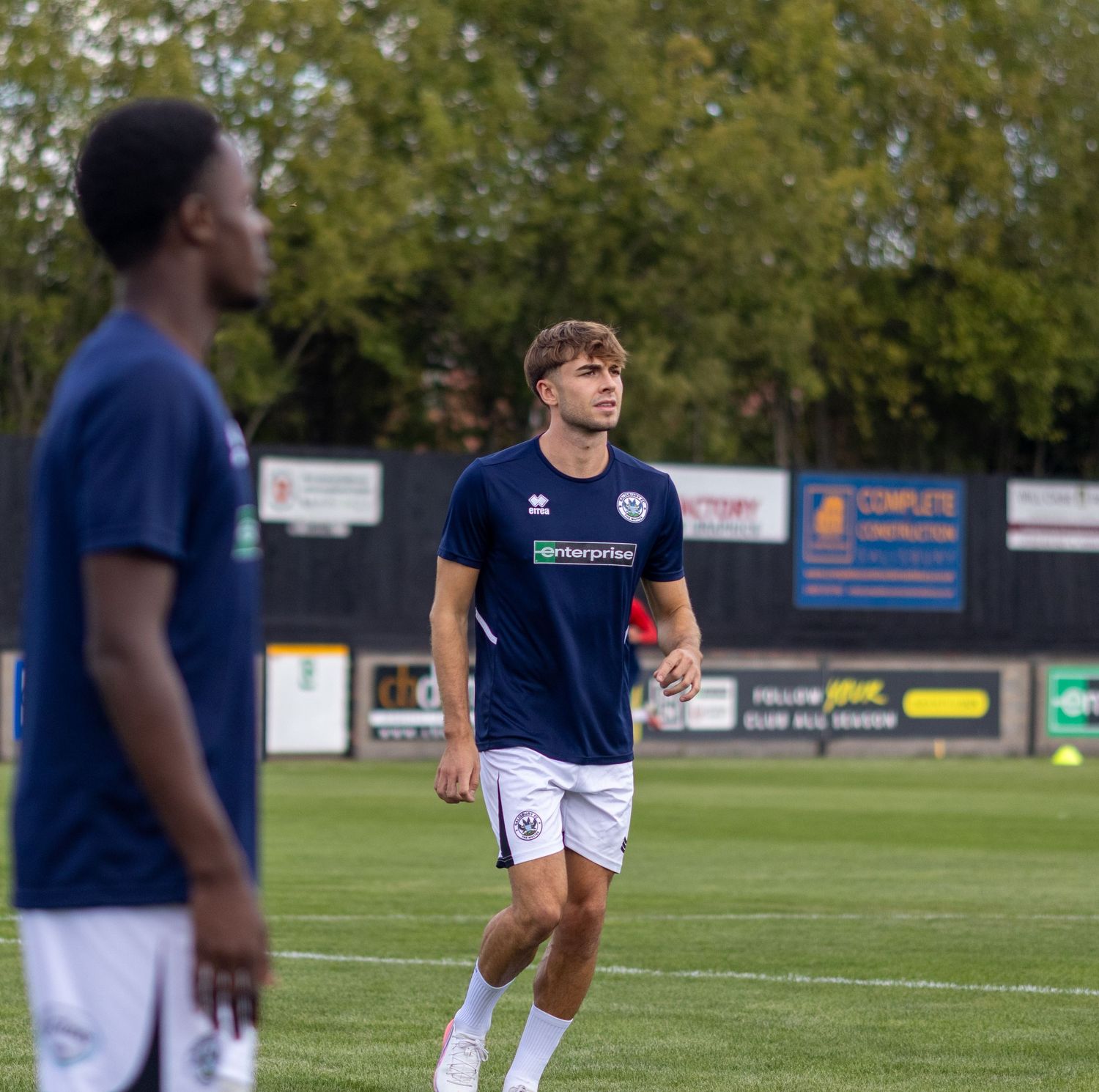 Two footballers in navy enterprise-sponsored kits during a pre-match warmup session on a grass football pitch.