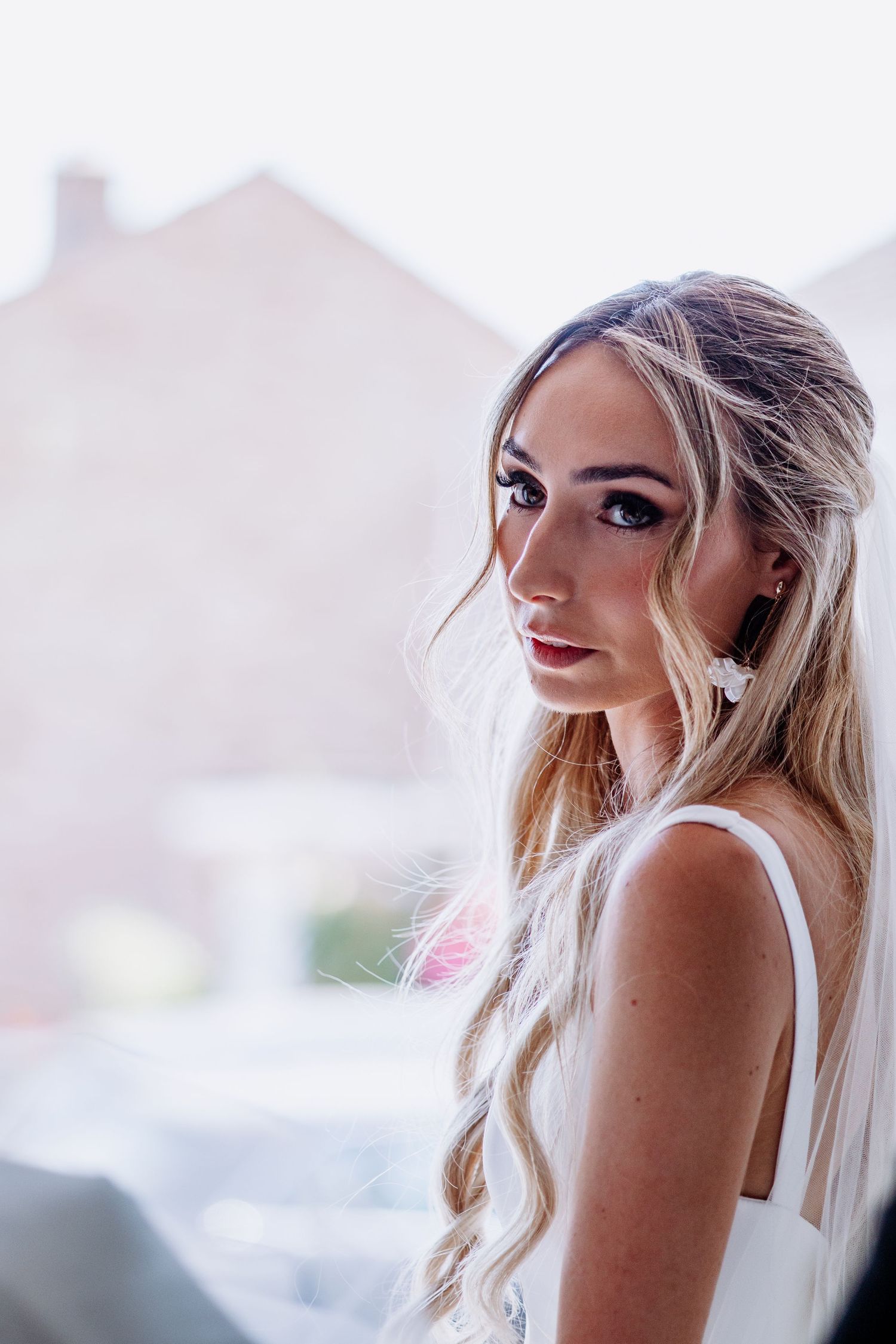 Elegant blonde bride with long wavy hair and pearl earrings poses in white dress near bright window.