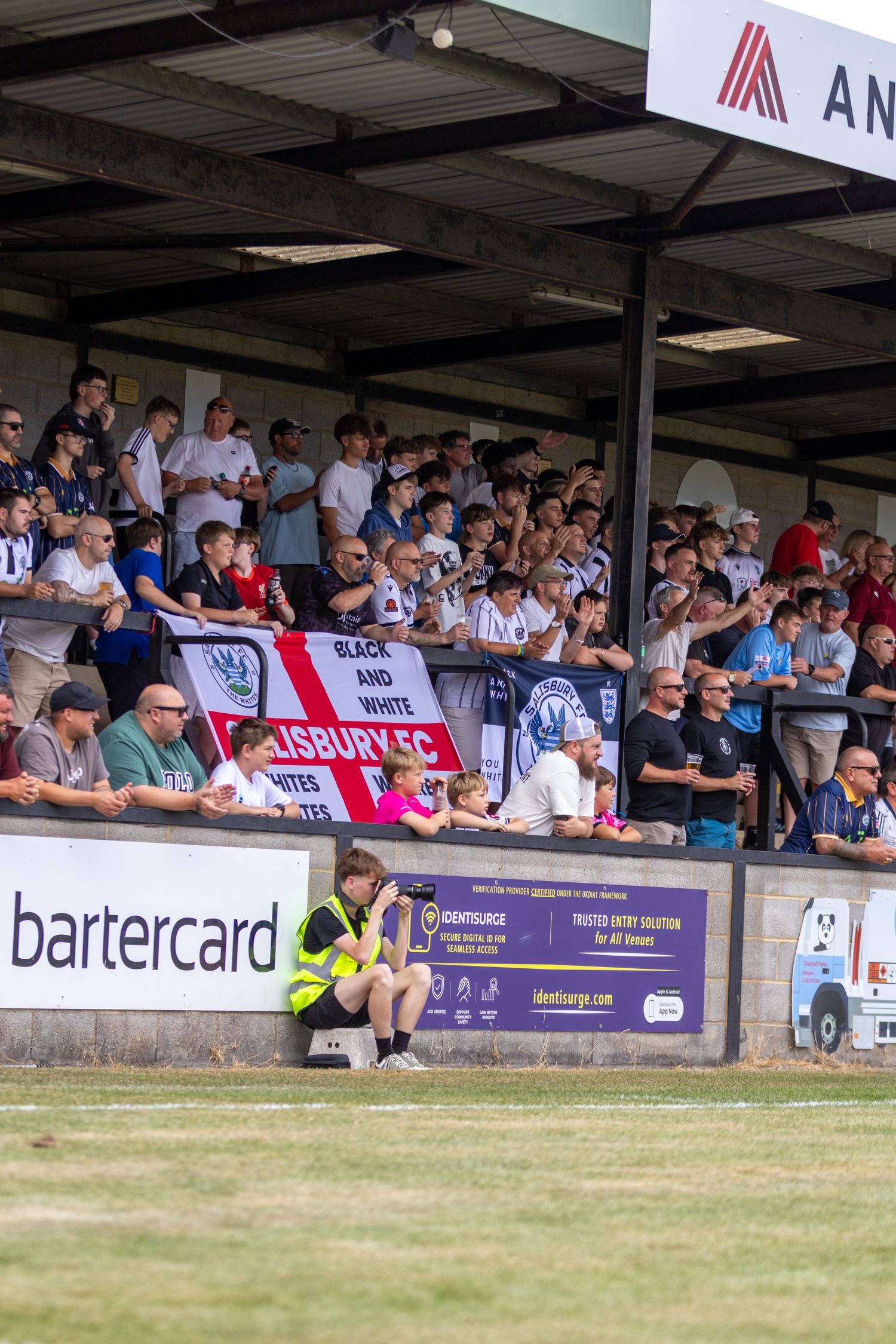 Salisbury FC supporters filling the covered stand with Black and White club banner on a sunny day.