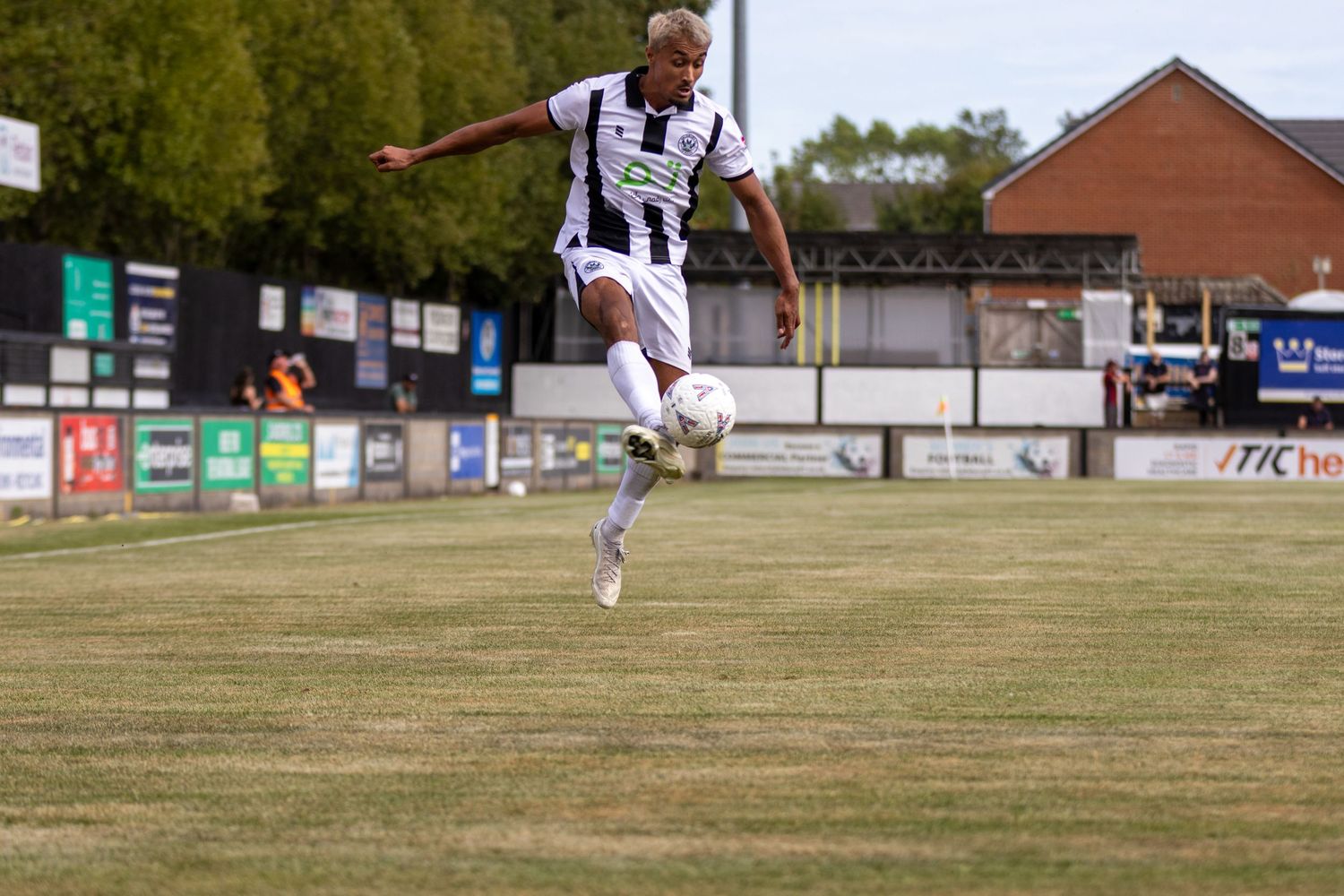 Athletic soccer player in black and white striped kit jumps to control ball on grass pitch near stadium.
