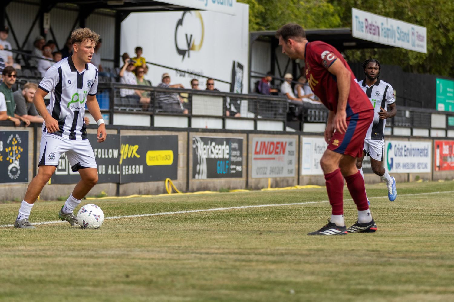 Two soccer players compete for the ball during a sunny outdoor match, one in black and white, one in red kit.