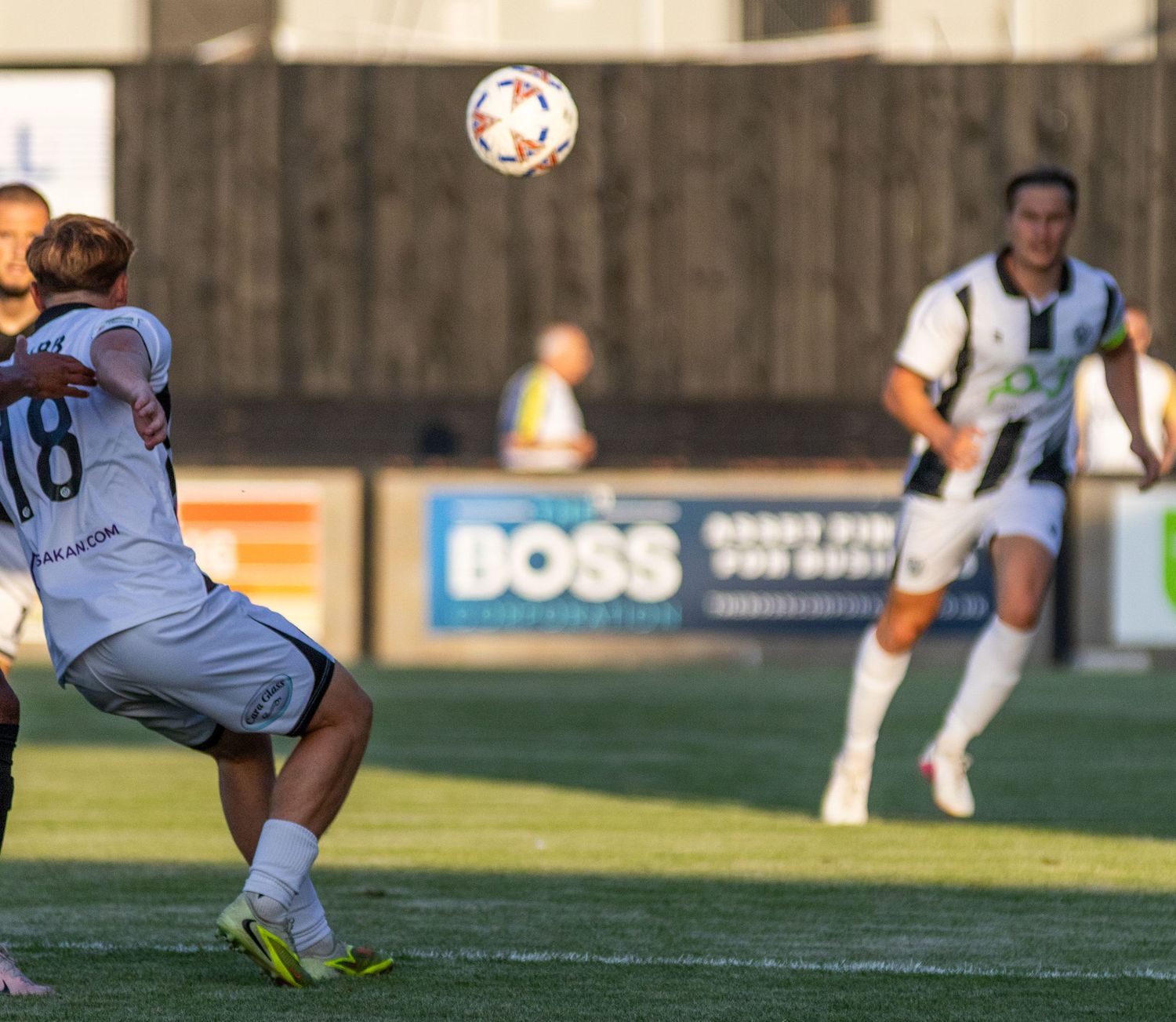 A footballer wearing number 18 controls the ball during a match with a BOSS advertising board behind.