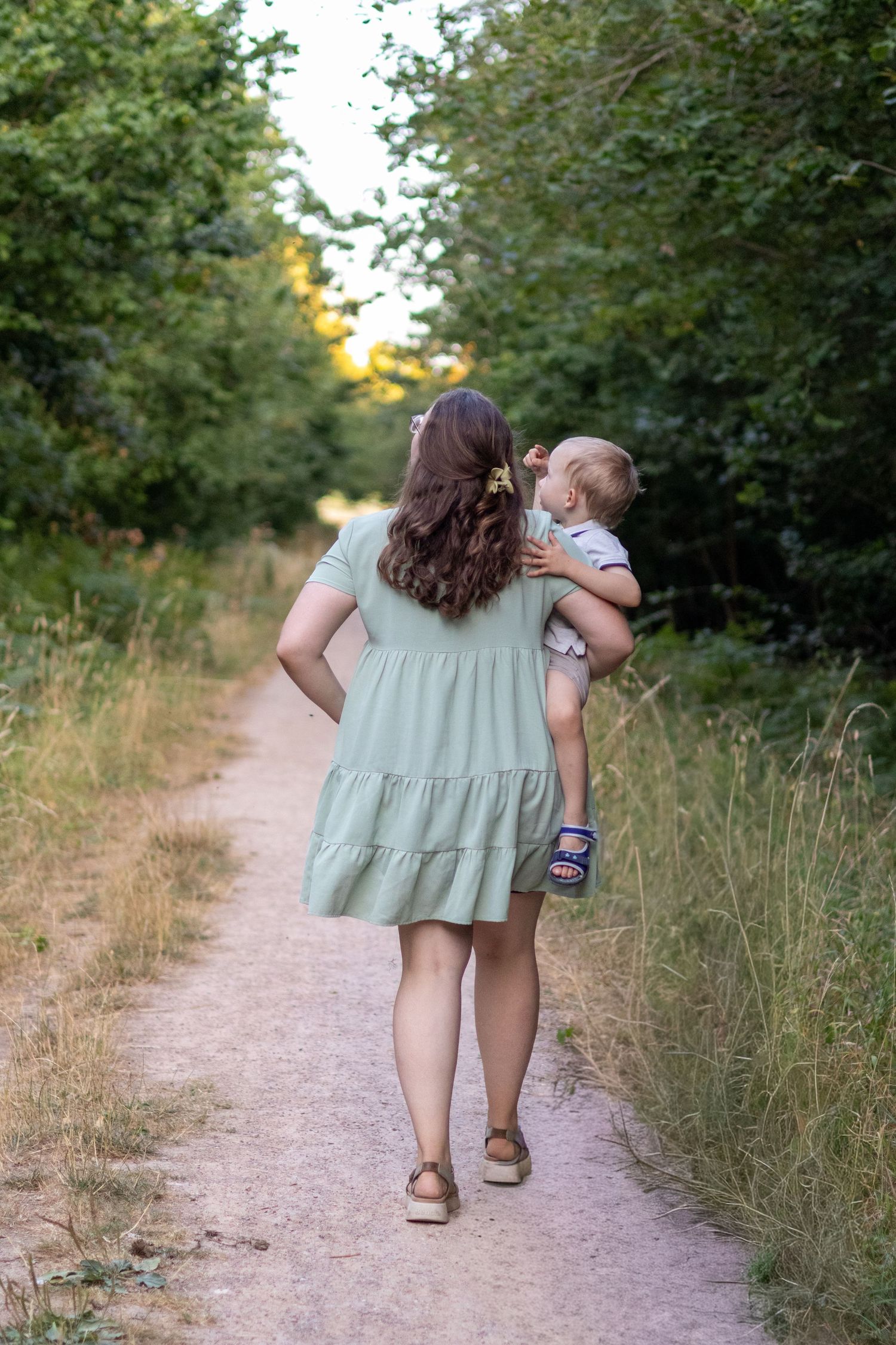 A mother in a sage green dress walks down a sunlit nature path while cradling her baby tenderly.