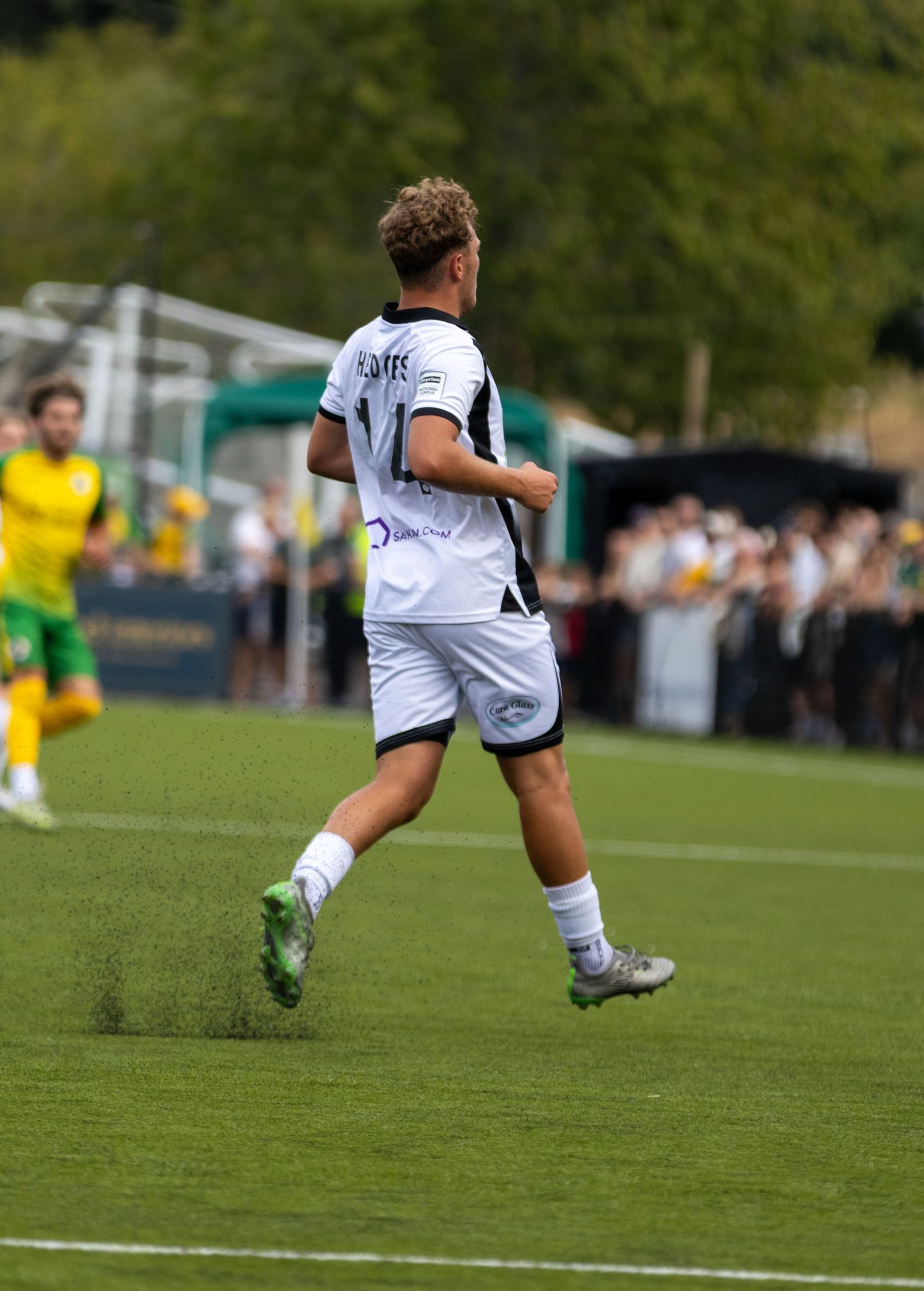 Footballer in white kit with curly hair runs across green pitch with yellow-kitted players in background.