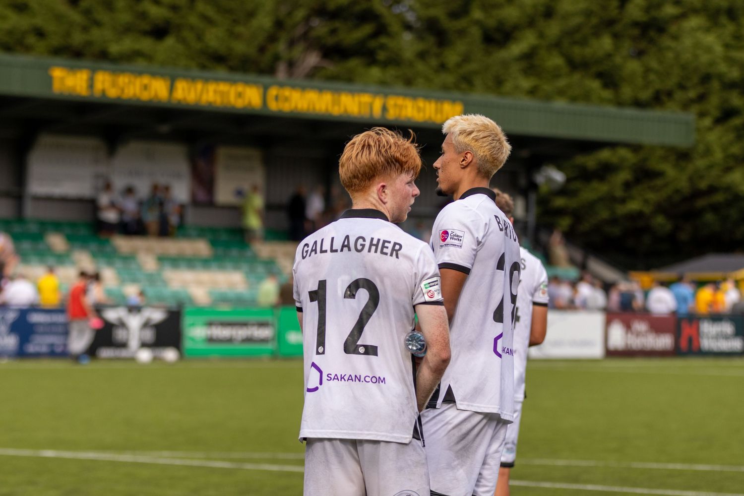 Two footballers in white kits, one wearing number 12 GALLAGHER, chat at a community stadium.