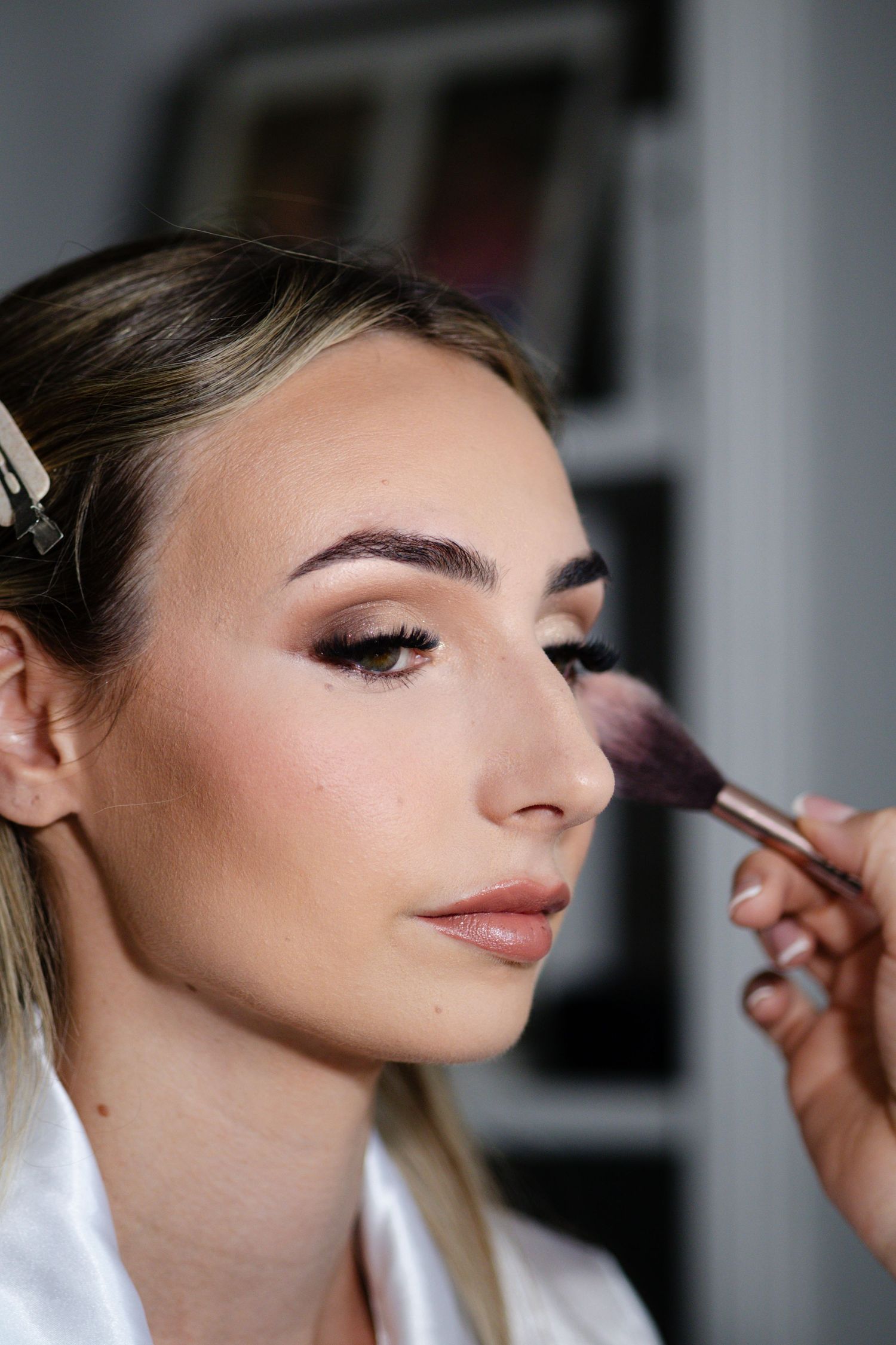 Professional makeup artist applying blush with a brush to a blonde woman in a white satin robe.