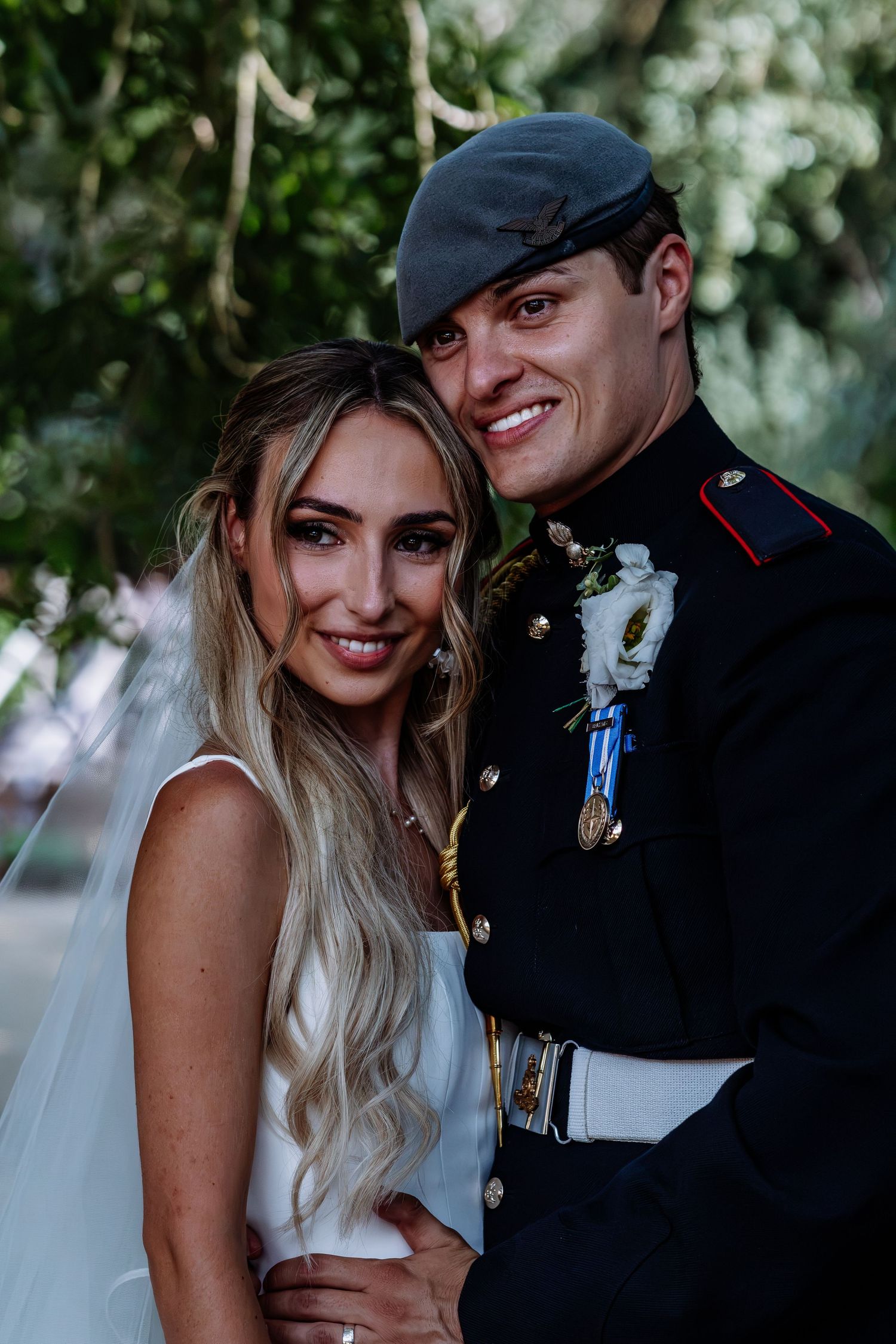 Smiling bride and groom in military dress uniform pose together outdoors.