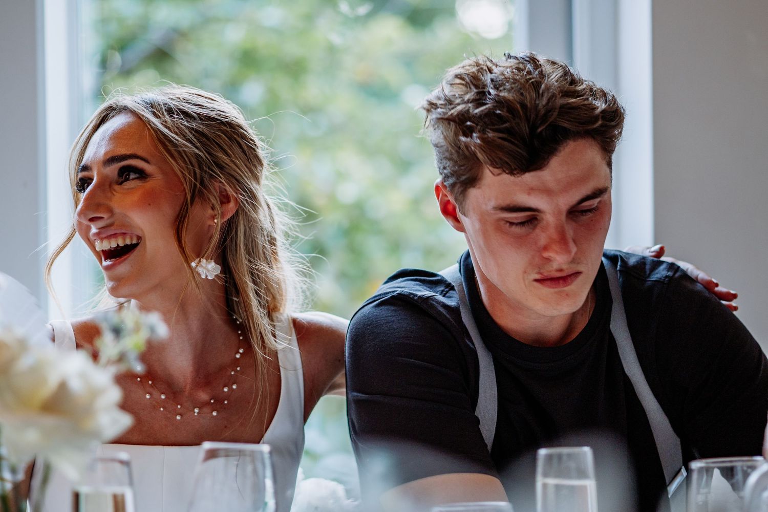 Bride laughing while groom smiles at wedding reception table with champagne glasses.