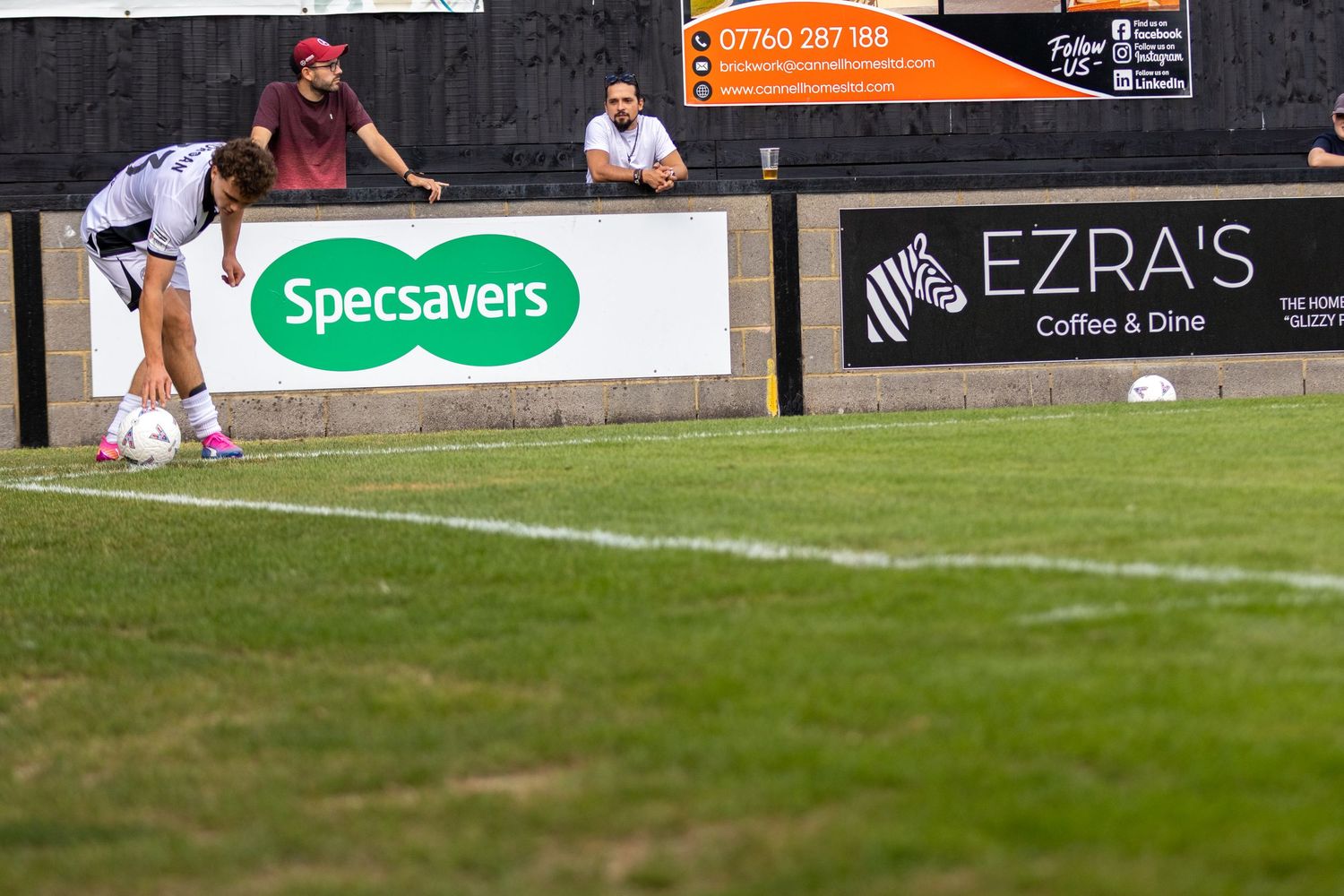 Soccer player kicking ball near sideline with Specsavers and Ezra's Coffee and Dine advertising boards.